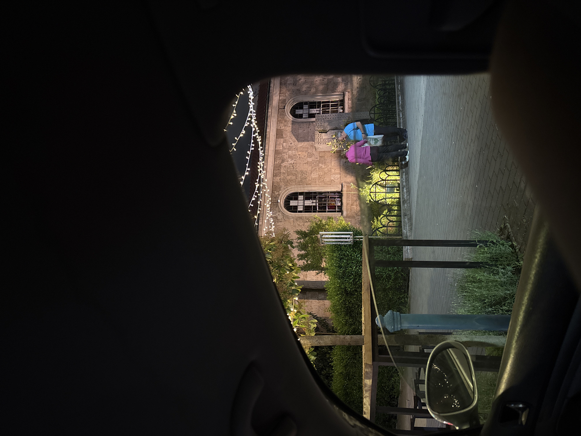 A nighttime view is presented from within a vehicle. Outside, two people are standing on a paved sidewalk alongside a building with illuminated windows. The building appears to be made of stone and is decorated with strings of lights. A short fence separates the sidewalk from a garden area. A street lamp and the car's side mirror are visible in the foreground. Based on the architectural style of the building and the lighting, the location is likely Burgas, Bulgaria.