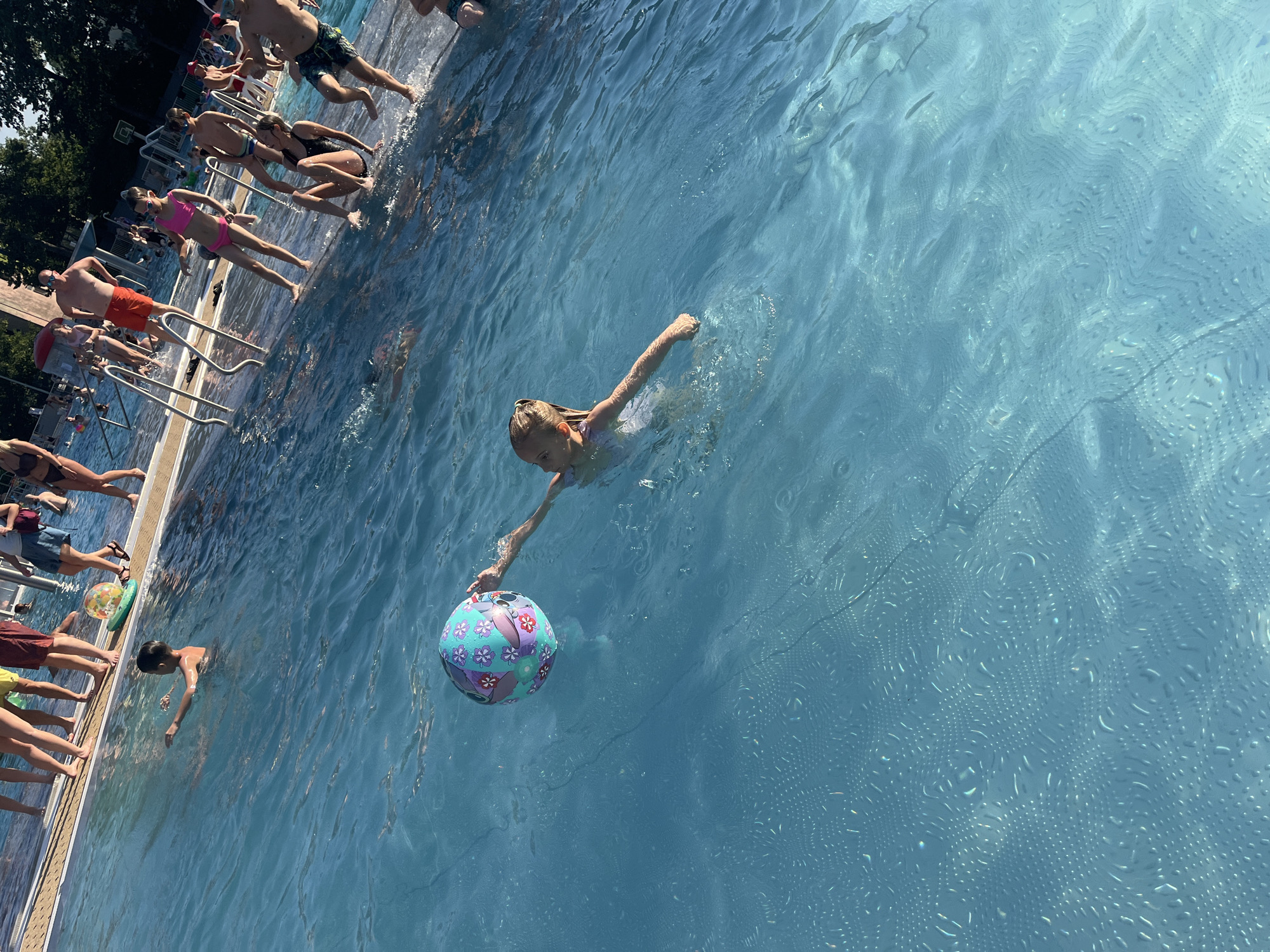 A group of people are gathered around and in a bright blue swimming pool. The shot is taken from an elevated angle, looking down into the water and towards the edge of the pool. Several individuals are standing or sitting on the pool deck, while others are swimming or wading in the water.
In the foreground, a young girl with blonde hair is swimming and holding a colorful inflatable ball. The ball is decorated with a floral pattern in shades of purple, pink, and blue. She is looking towards the ball and appears to be engaged in play. Other swimmers are visible in the background, some near the pool's edge and others further out in the water. The pool appears to be outdoors, with sunlight reflecting off the water's surface, creating a shimmering effect. Trees and some structures are visible in the background on the left side, suggesting a park or recreational area. The overall atmosphere appears to be relaxed and summery, with people enjoying a day at the pool. The time of day is likely midday due to the bright sunlight and strong shadows. No text is visible in the image.
