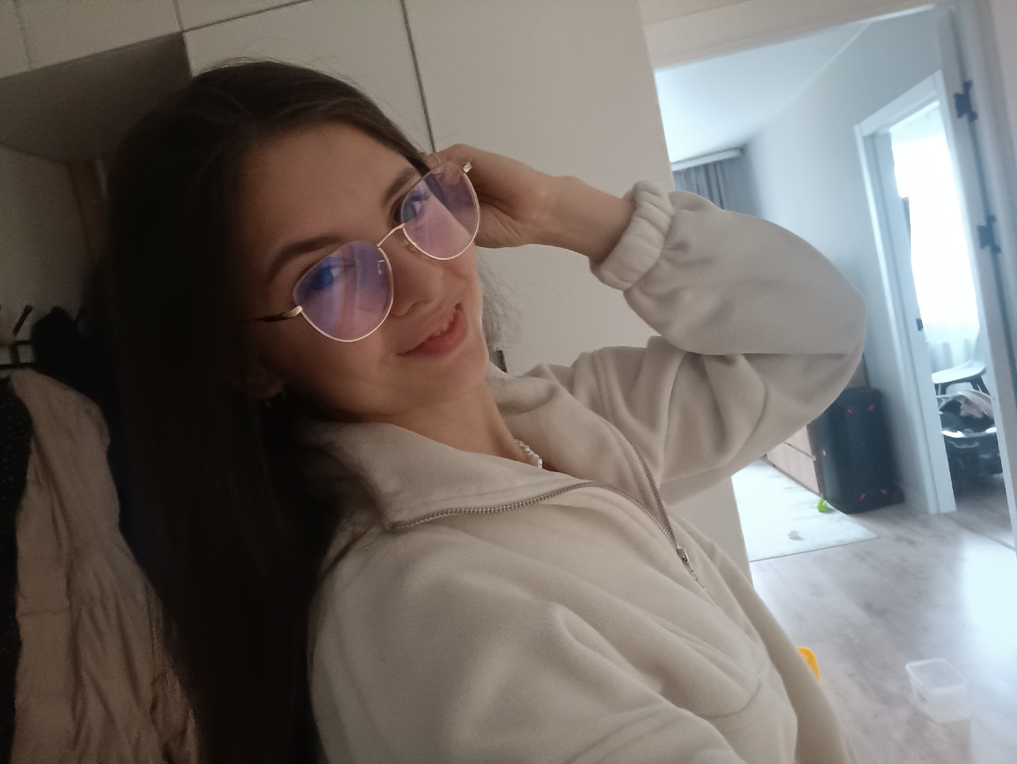 A young woman with long dark hair and light skin is posing for a selfie indoors. She is wearing round, gold-rimmed glasses and a light-colored, zip-up sweatshirt. Her right hand is raised near her glasses.
Behind her is an open doorway leading to another room with light walls and a window. A gray curtain hangs by the window. A black, cylindrical object with red dots is visible in the room beyond, along with a white rug, light wood furniture, and a white chair. A clear plastic container is on the floor near the woman. Part of a closet with neutral-toned garments can be seen on the left side of the frame.
