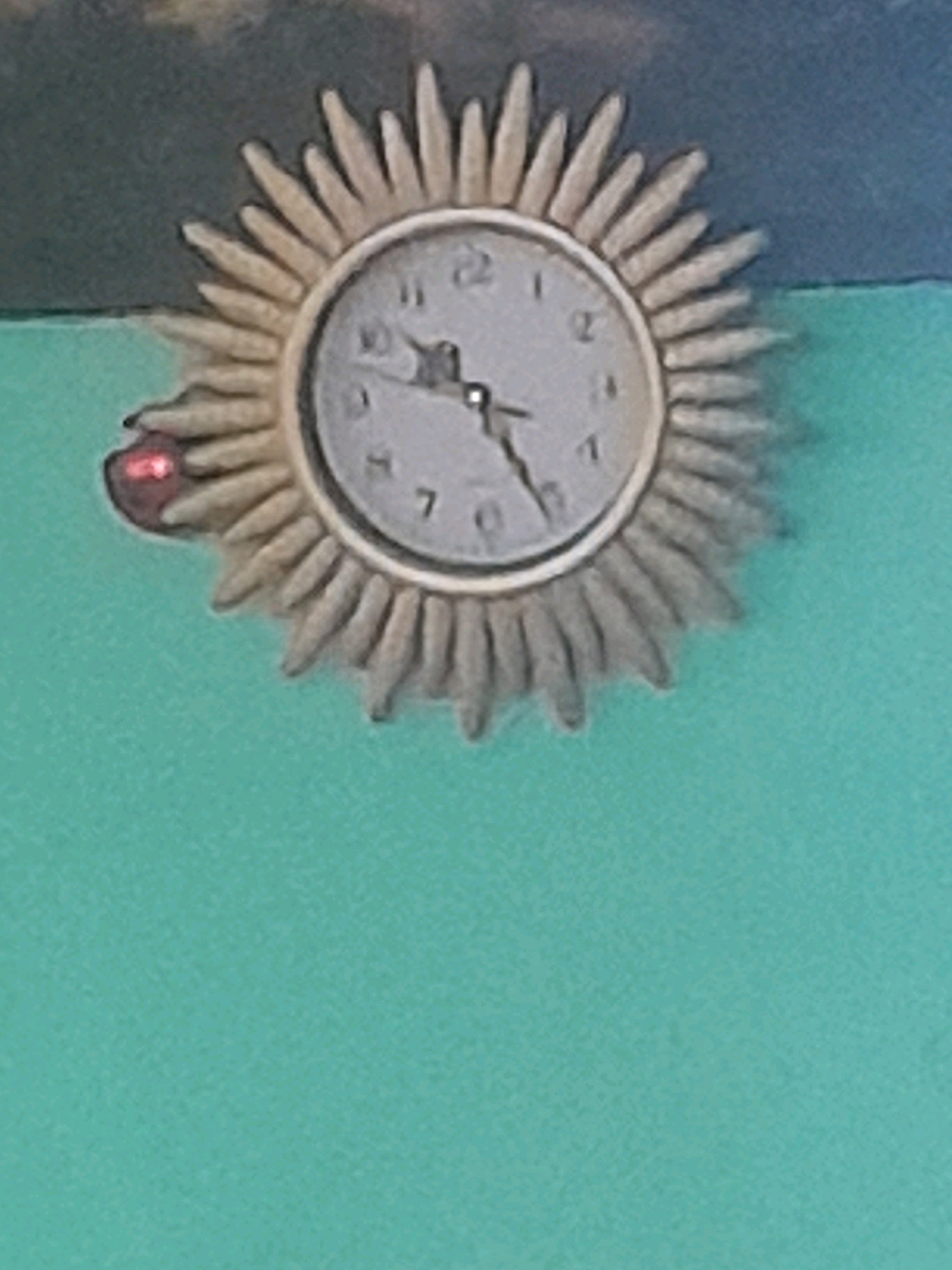 A close-up shot captures a sunburst clock mounted on a teal-colored wall. A small red object, possibly a sticker or a small decoration, is affixed to one of the clock's rays on the left side. The clock face features Roman numerals, and its hands are partially visible. The background shows a muted, textured pattern in darker tones, suggesting a wall or perhaps a mural. The image is slightly out of focus, giving it a soft, diffused appearance.