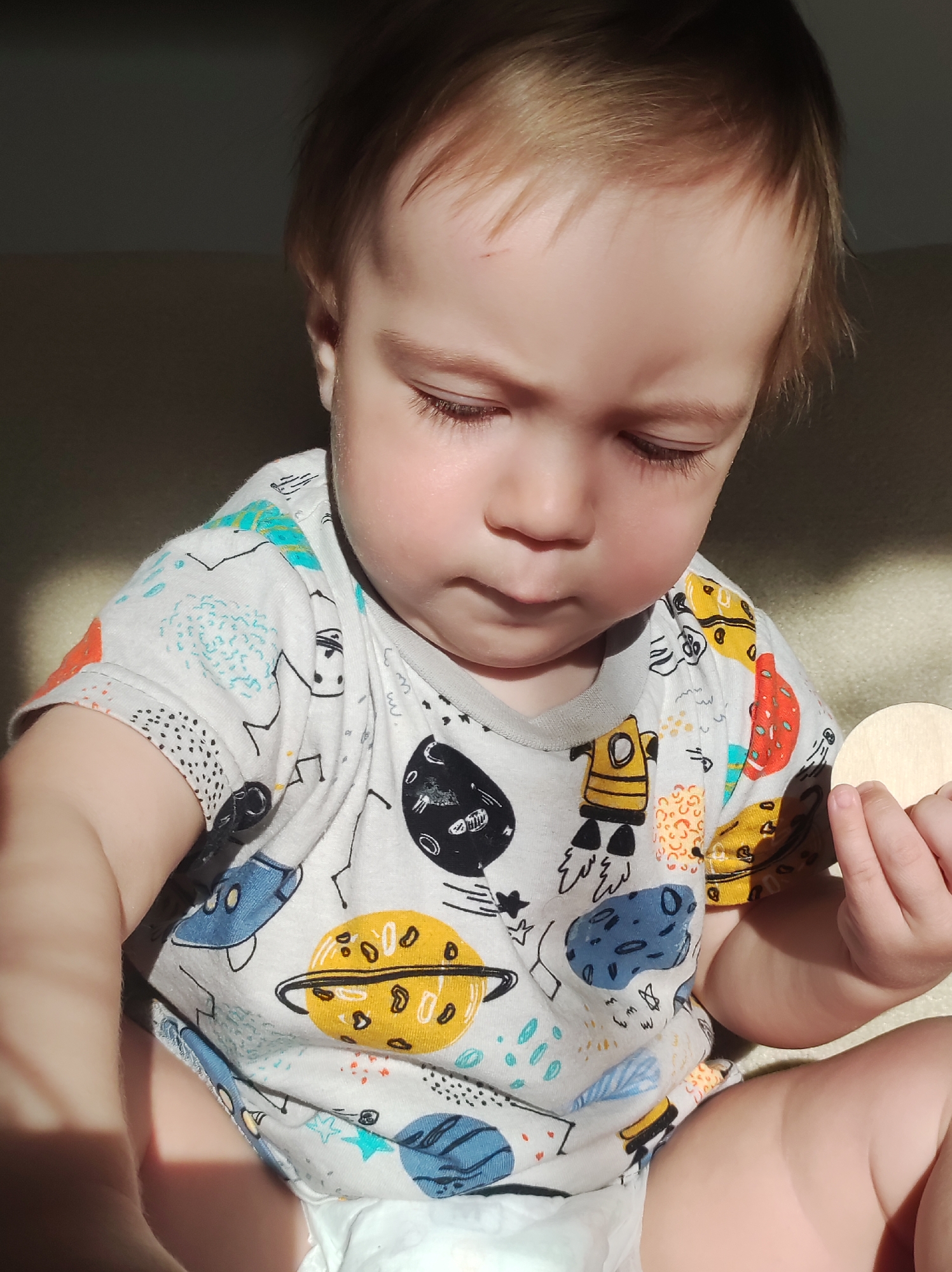 A baby is sitting down, wearing a white t-shirt with colorful planet and space-themed prints. The baby is wearing a diaper. The baby is holding a small, round, wooden object in their right hand. The setting appears to be indoors. There is no information to indicate the location is Фоки, Russia.