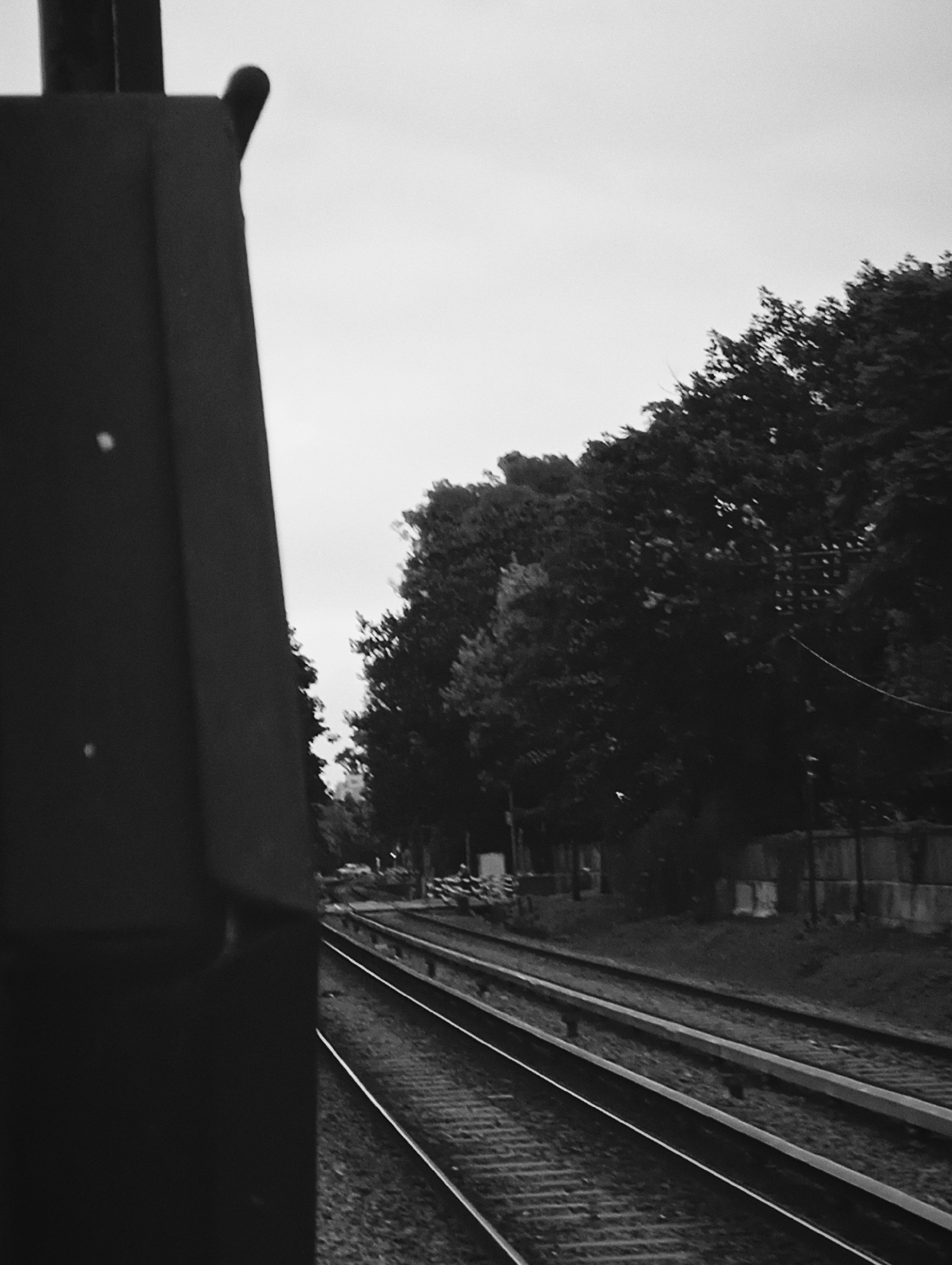 The black and white image depicts a view from a train, looking out over railway tracks. A dark, partially visible structure is on the left side of the frame. Parallel railway tracks run into the distance, flanked by trees and a fence on the right. The sky is overcast. The location is likely Buenos Aires, Argentina, based on the urban railway infrastructure and architectural style, although specific landmarks are not visible to confirm.