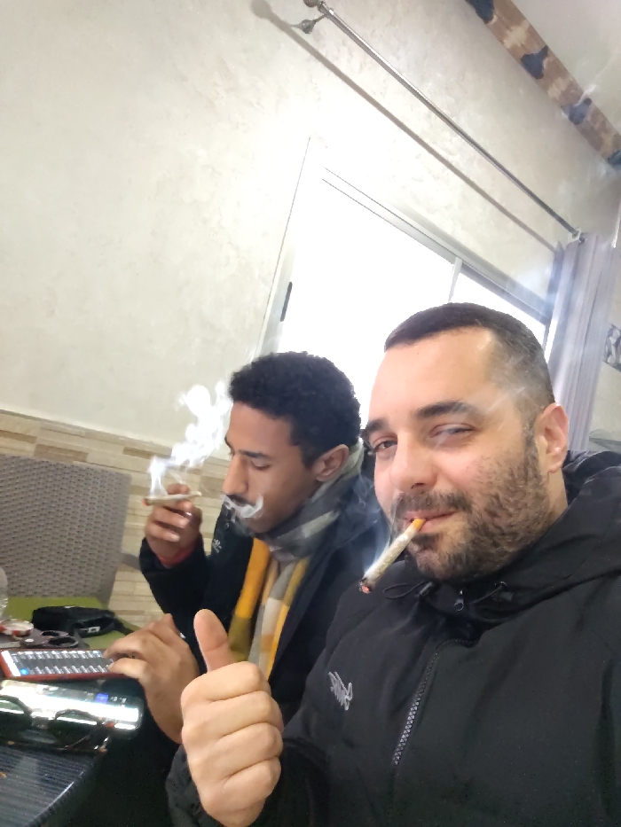 Two men are sitting indoors, each holding a lit marijuana cigarette. The man in the foreground wears a black jacket and gives a thumbs up. The other man has curly hair, a scarf, and smoke billowing from his joint. A table cluttered with personal items sits between them. The background features a window and textured walls.