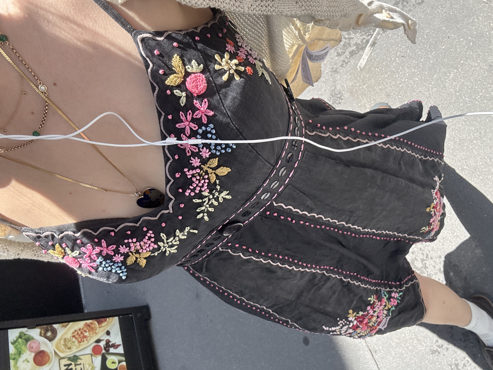 The image shows a close-up, top-down view of a person from the chest down, wearing a black dress with colorful floral embroidery. The dress has thin spaghetti straps and a scalloped neckline. The embroidery features pink, blue, yellow, and white flowers, along with green leaves. The waistline of the dress is cinched with a decorative band of small oval shapes and a button. Pink stitching accents the edges of the dress panels. A cream-colored, knitted cardigan is draped over the person's shoulders. White wired earbuds are visible, with the wire running down the front of the dress. The person is wearing multiple necklaces, including a gold chain with a dark heart-shaped pendant and another with green beads and a diamond. The background appears to be an outdoor urban setting, with a grey sidewalk and a shadow cast by the person. To the left, a sign with food images is partially visible, suggesting a restaurant or food establishment. The scene is brightly lit by sunlight. The setting is New York City, USA.