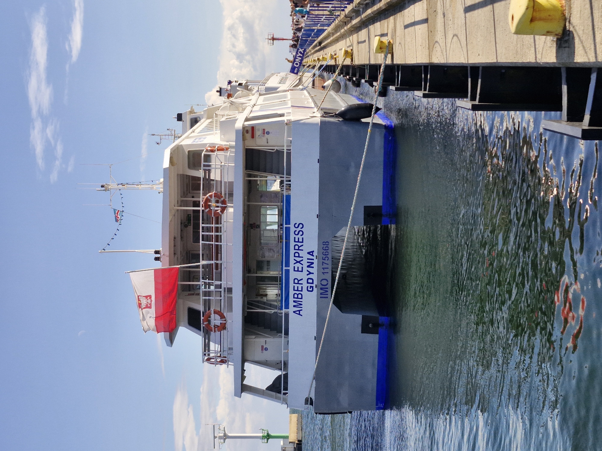 The image shows the rear view of a catamaran ferry docked at a pier in Hel, Poland. The ferry is named "AMBER EXPRESS GDYNIA" and has the IMO number 1175668 displayed on its hull. The Polish flag is prominently displayed on the deck of the ferry. Several life preservers are visible on the railings. The water is calm, with reflections of the ferry and the sky visible. The pier is made of concrete with yellow bollards and black mooring posts. In the background, people are walking along the pier, and a red lighthouse is visible. A green and white lighthouse is also present in the distance to the left. The sky is blue with scattered clouds.
