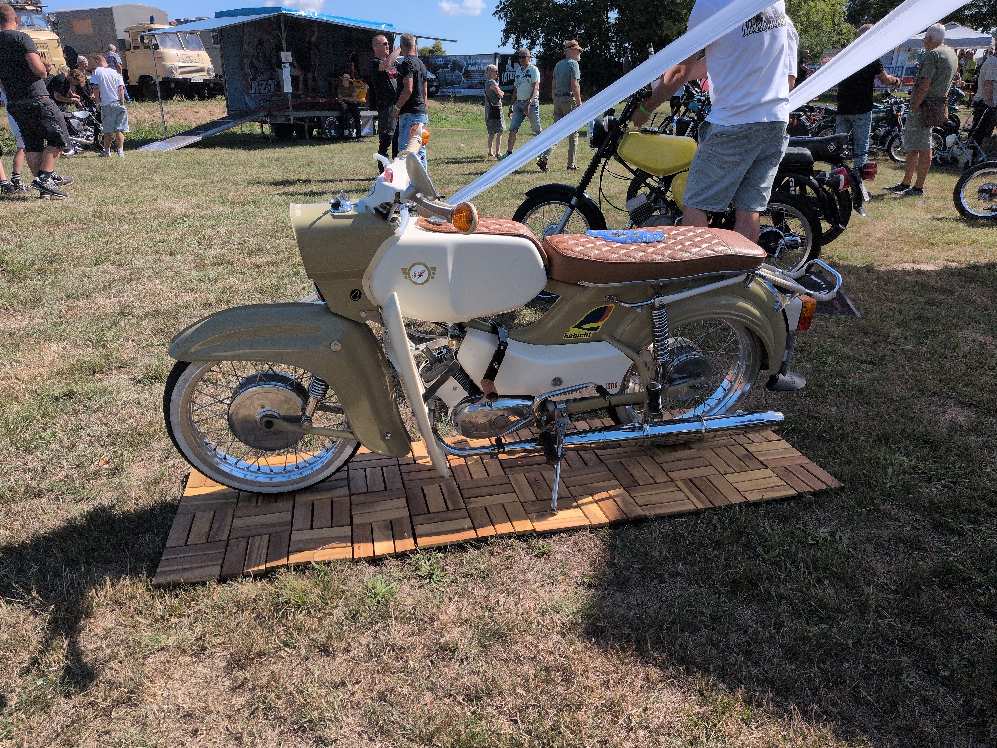 This image captures an outdoor event, likely a gathering or festival, in the city of Hohenferchesar, Germany, on a sunny day. In the foreground, a vintage moped, predominantly olive green and cream with a brown diamond-stitched seat, is displayed on a wooden slatted platform. The moped features chrome accents and a visible kickstand. Behind it, a yellow moped is partially visible.
Several people are present in the background, mingling and observing. Some are standing near a blue and white tent-like structure with "NZ" visible on its side. Others are near various vehicles, including an older tan truck with a blue awning and a lighter colored truck with a dark roof. The ground is covered in dry grass, and the scene is bathed in sunlight, casting shadows from the objects and people. The overall atmosphere suggests a casual outdoor event focused on vintage vehicles.