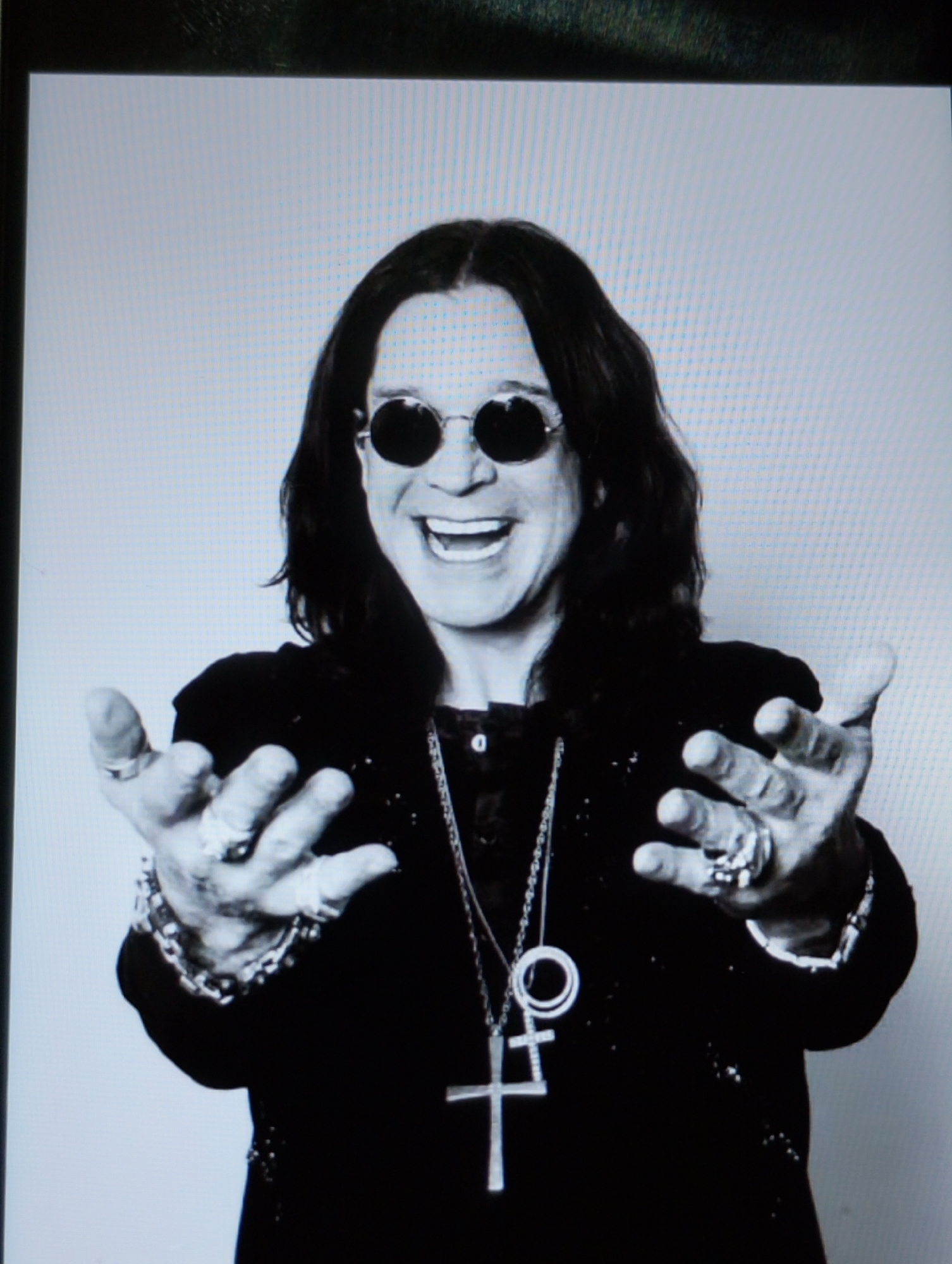 A black and white medium shot captures a man with dark, wavy hair, wearing large, round sunglasses. He is smiling widely, with his mouth open, and has his hands outstretched towards the viewer. His left hand displays several rings on its fingers and a beaded bracelet on his wrist. He is wearing a dark, possibly black, jacket over a dark shirt. A prominent silver necklace with a cross pendant and a circular charm hangs around his neck. The background is a plain, light gray. The overall impression is of a posed, candid portrait.