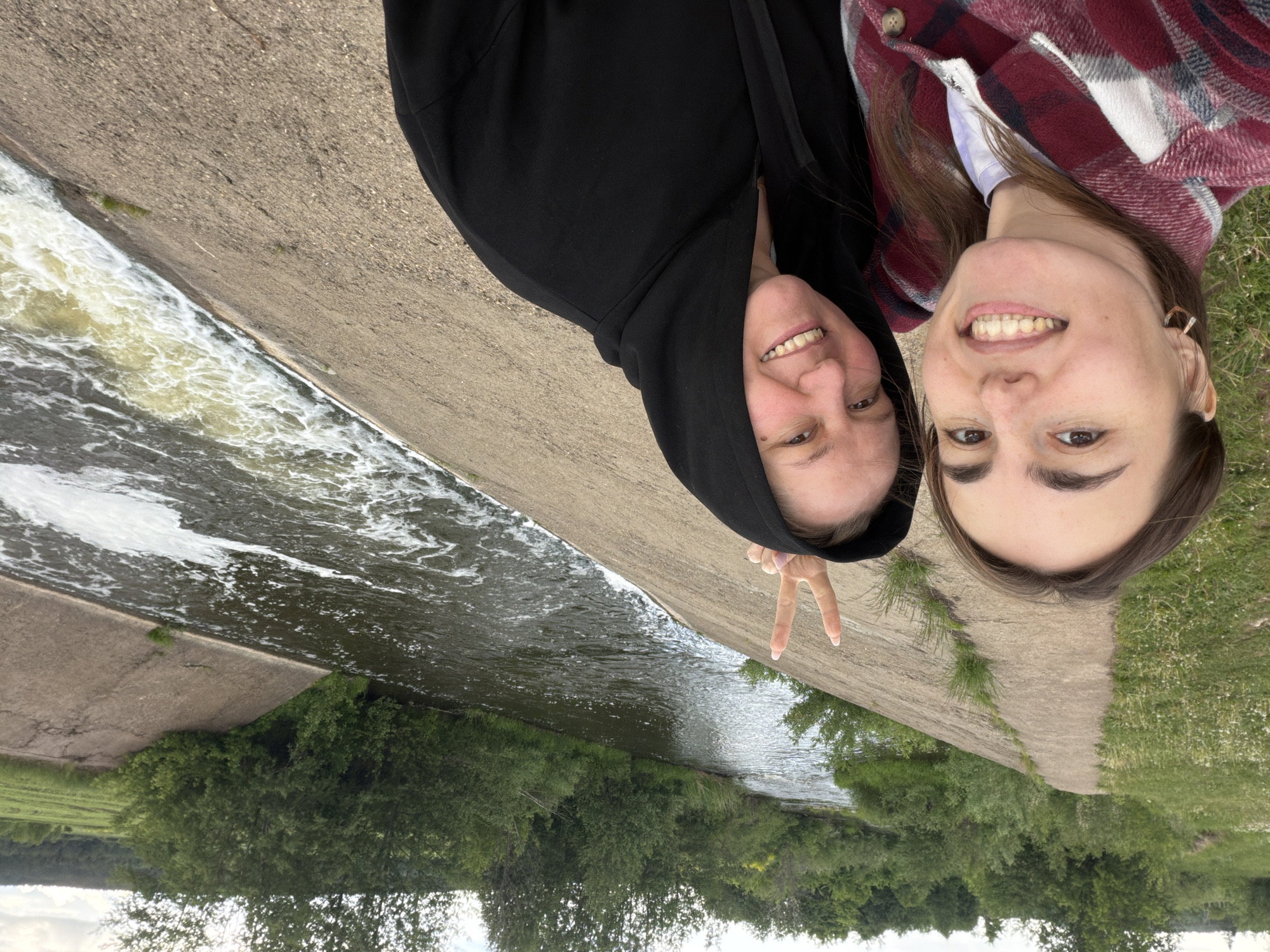 Two light-skinned women are positioned close together, smiling at the camera. The woman on the left wears a black hooded sweatshirt and holds up two fingers in a peace sign. The woman on the right wears a plaid shirt over a white top. They are standing on a concrete edge next to flowing water. Below them is a wooded area. The location is Nizhnii Vishur, Russia.