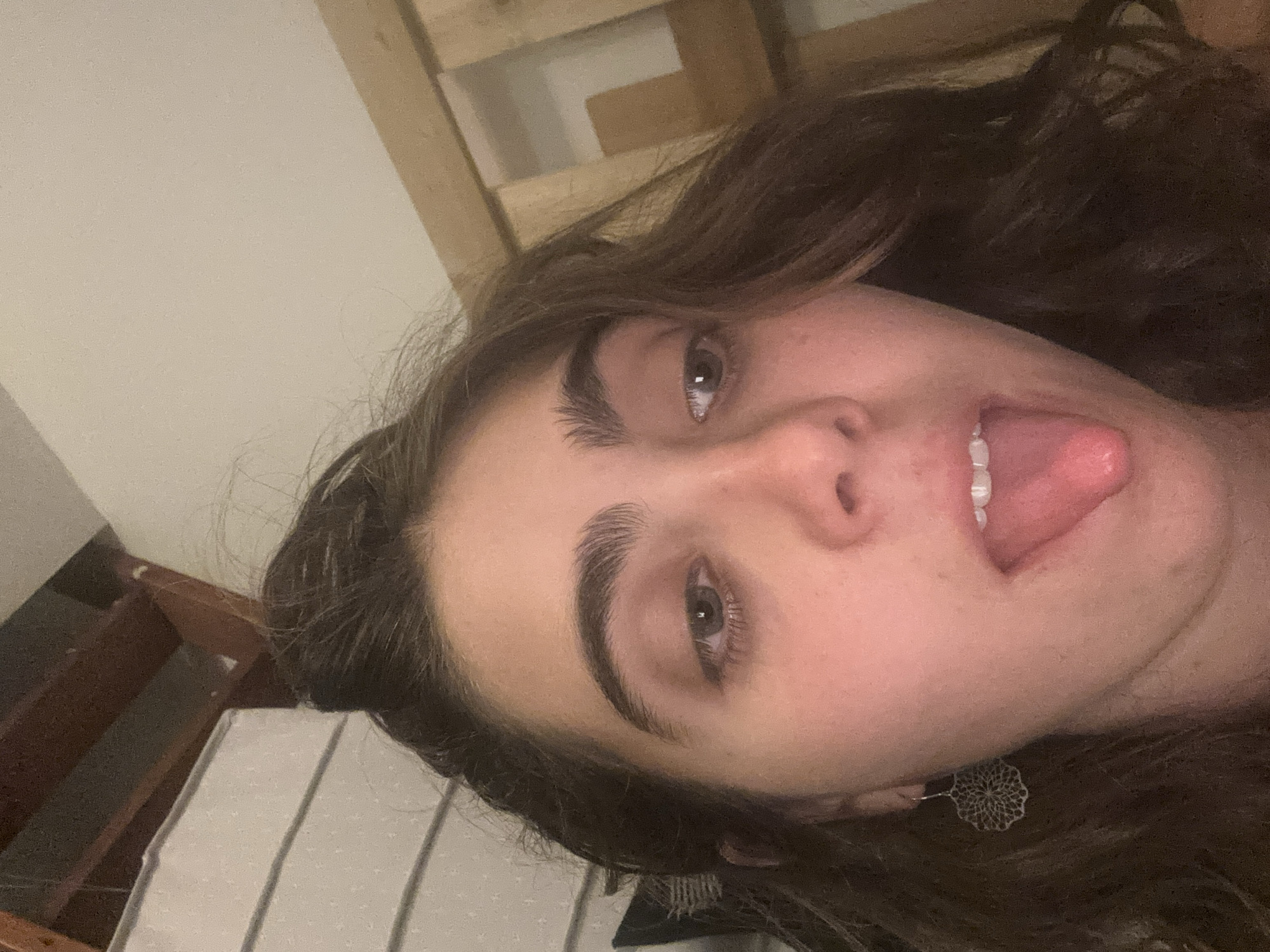 A selfie of a fair-skinned person with long, dark brown hair. The person is sticking their tongue out. They have dark eyebrows and light-colored eyes. A silver earring is visible on their left ear. The background is blurry and includes what appears to be a wooden structure and a white striped surface. The image does not depict any recognizable locations in Tel-Aviv, Israel.