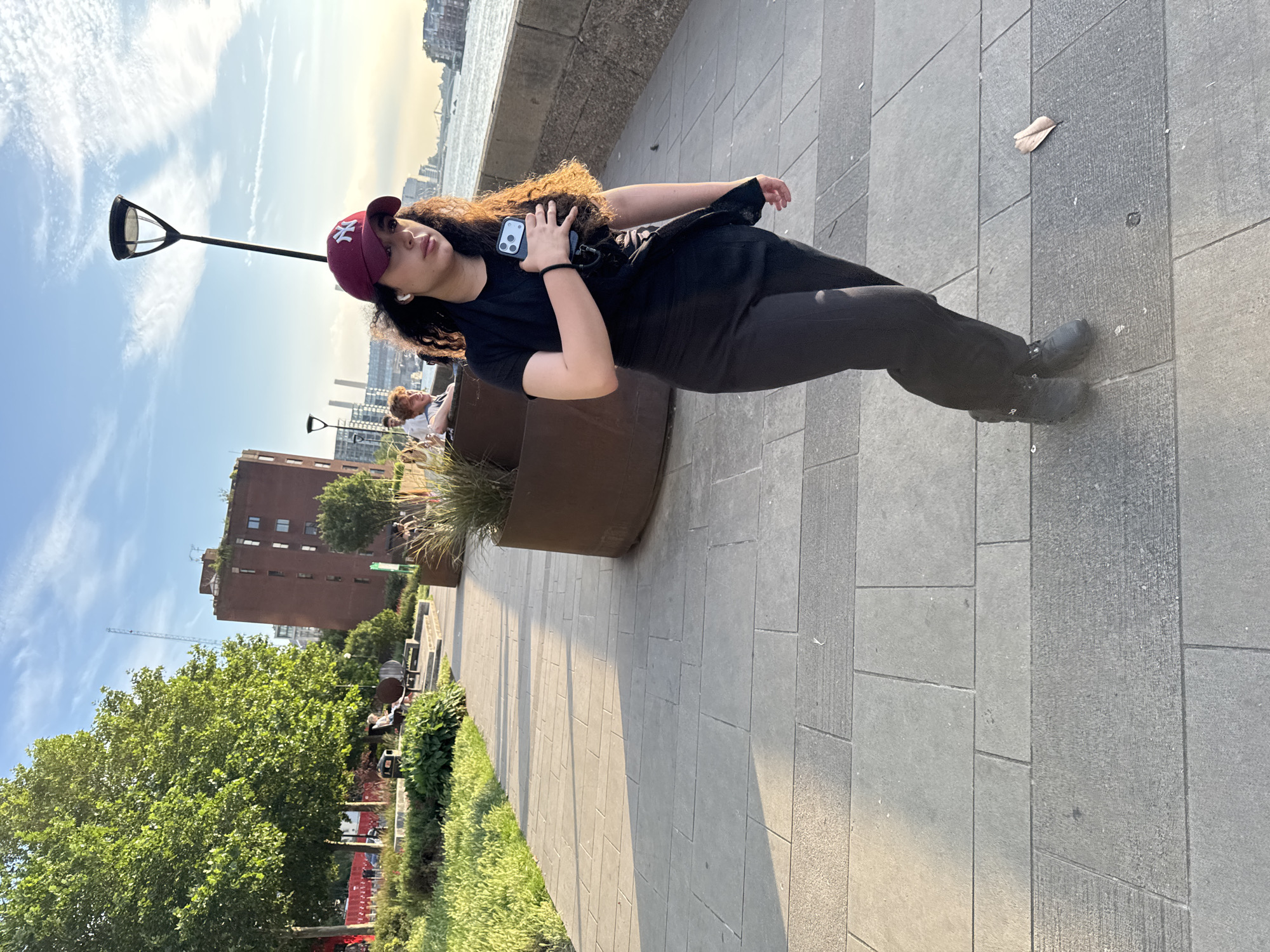 A young woman in a black t-shirt, black pants, and a maroon baseball cap with a white "NY" logo walks on a paved walkway next to a body of water. She is holding a smartphone in her right hand, and wearing white earbuds. The walkway is lined with planters containing dry grasses. In the background, there are trees, a brick building, and other people sitting on benches. The scene appears to be on the South Bank of the River Thames in London, United Kingdom, given the architectural style of the buildings and the presence of the river. The sun is low in the sky, casting long shadows.