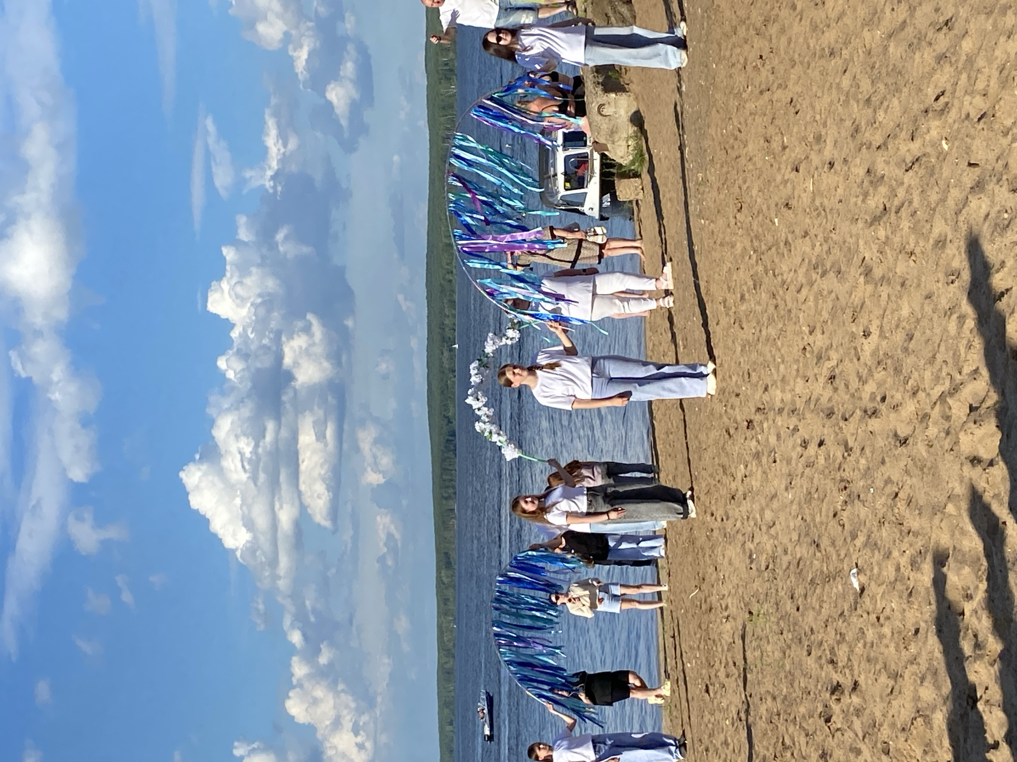 A group of people are gathered on a sandy beach by a body of water, likely a lake or river, under a bright blue sky with scattered clouds. The sun is shining, suggesting it is daytime. The people are arranged in a line, with some standing and others partially visible, facing towards the water. They are mostly dressed in light-colored clothing, such as white tops and blue jeans, giving a casual and perhaps festive atmosphere.
A large, semi-circular decorative arch adorned with blue and purple ribbons is a focal point of the scene, held by some of the individuals. White floral arrangements are also visible, possibly part of the same decoration or a separate element. Behind the people and the arch, the water stretches out, with a hint of green shoreline in the distance. A small boat is visible on the water. The sandy ground has footprints and some scattered debris. The overall impression is of an outdoor event or gathering, possibly a celebration or performance, taking place on a sunny day.