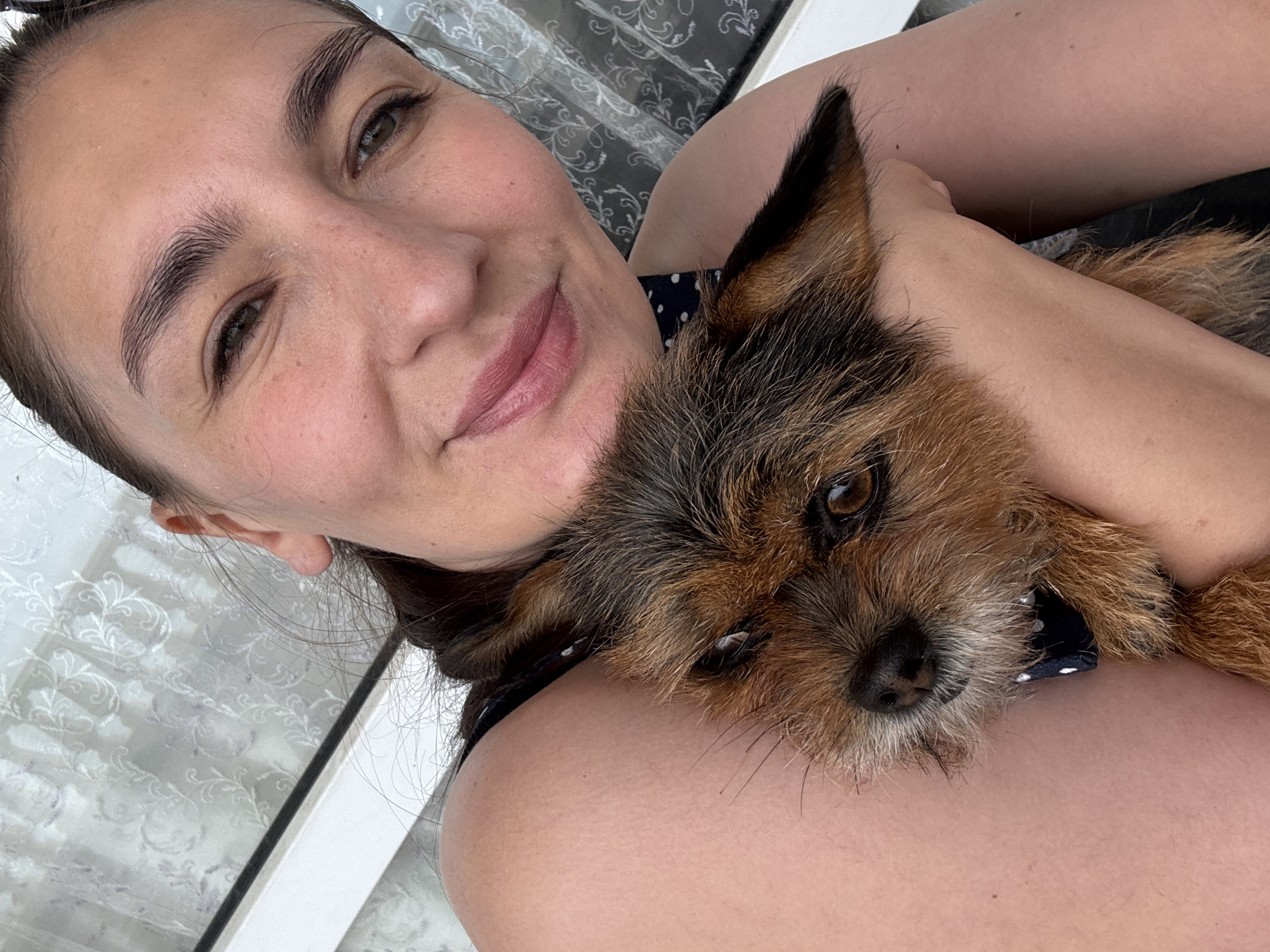 A selfie depicts a smiling woman holding a small dog against her chest. The woman has brown hair pulled back, olive skin, and brown eyes. She is wearing a dark top with white polka dots. The dog has brown and tan fur and appears to be a terrier mix. The background is blurred, but shows a window with a patterned curtain and a white frame. There is no indication that this image was taken in Sochi, Russia.