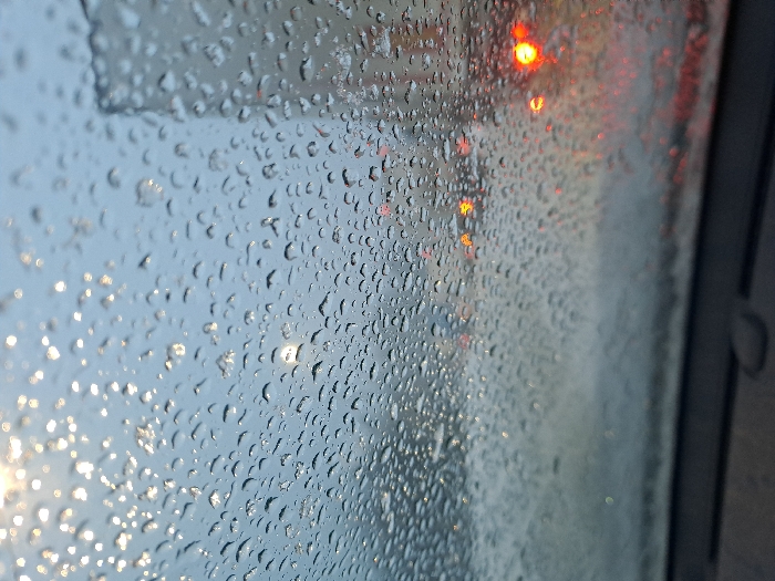 Raindrops cover a window, blurring the view beyond. Lights appear distorted through the water droplets, creating bokeh effects. The scene suggests wet weather conditions, with a combination of natural light and artificial light sources visible. The close-up view emphasizes the texture of the raindrops on the glass surface.
