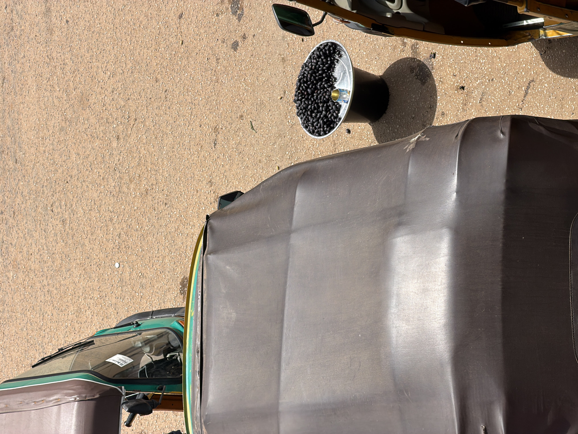 The image captures an overhead view of an outdoor scene, likely in Bukuru, Nigeria. The primary subjects are two auto-rickshaws, commonly known as "keke napeps." One auto-rickshaw is predominantly green and brown, with its windshield and front partially visible. The other auto-rickshaw, mostly obscured by its canopy, appears to be dark brown or black.
In between the two vehicles, a metallic, conical container is placed on the ground. This container is filled with numerous small, dark, berry-like objects, which appear to be olives. The ground is a textured, light brown asphalt surface, typical of a road or paved area. A shadow cast by an unseen object, possibly the container or another vehicle, is visible on the ground to the left of the container. A side mirror from a vehicle is also visible in the upper right portion of the image.
The lighting suggests it is daytime, with the sun casting distinct shadows. The scene appears to be a moment of pause or activity related to the sale or transport of the olives. There are no people clearly visible in the frame, but the presence of the auto-rickshaws implies a setting where transportation and possibly commerce are taking place. No text is discernible in the image, except possibly a faint label on the windshield of the green auto-rickshaw.