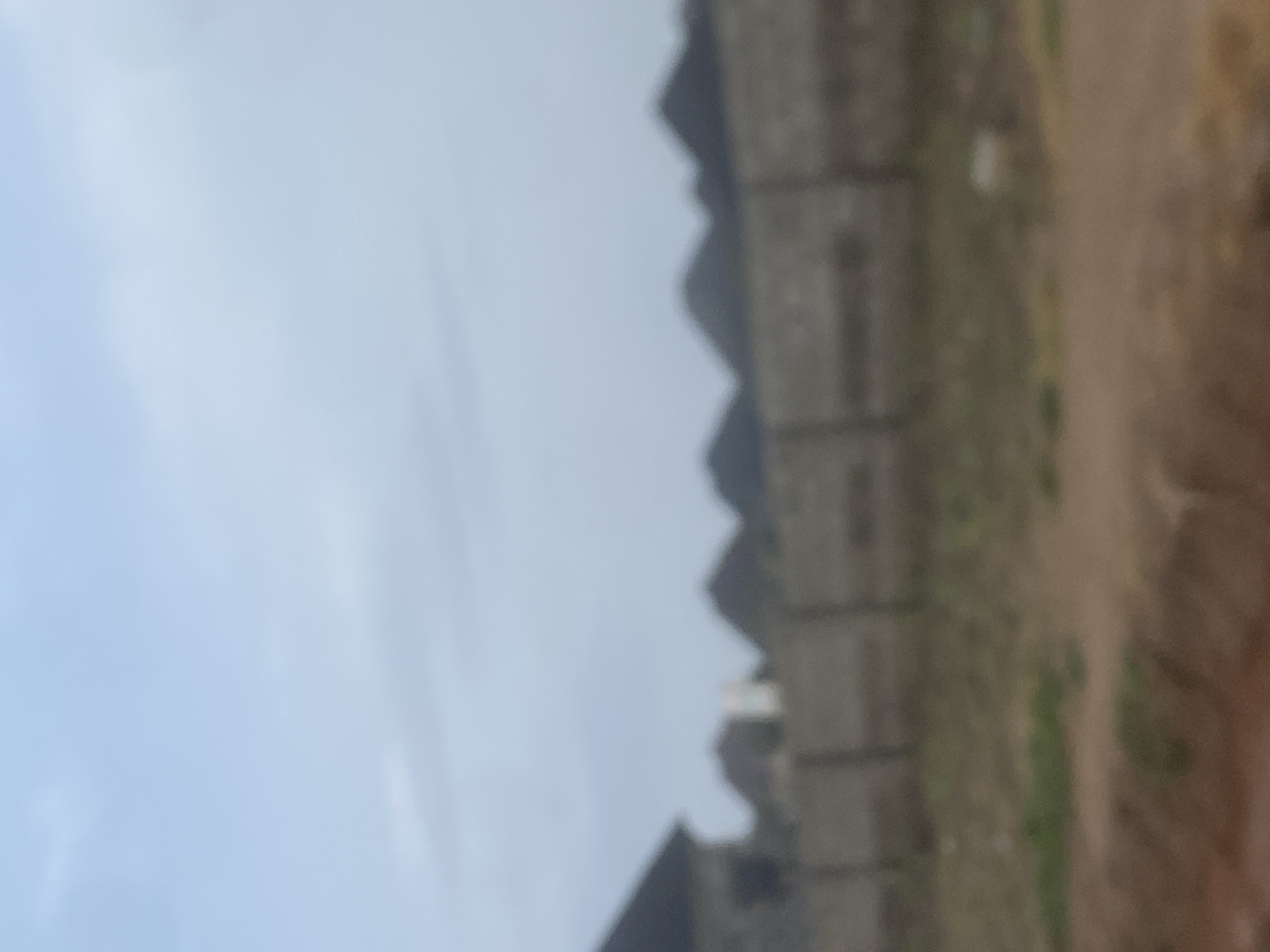 The image shows a blurred view of a street in Iddo Tudunwada, Nigeria. A low, textured wall runs horizontally across the mid-section of the frame, with what appear to be the dark, angular roofs of buildings peeking over it in the background. The sky above is a hazy, light blue, with some faint clouds. The foreground consists of a rough, uneven ground with patches of dry grass and some scattered debris. The overall impression is one of an overcast day in a developing area.
