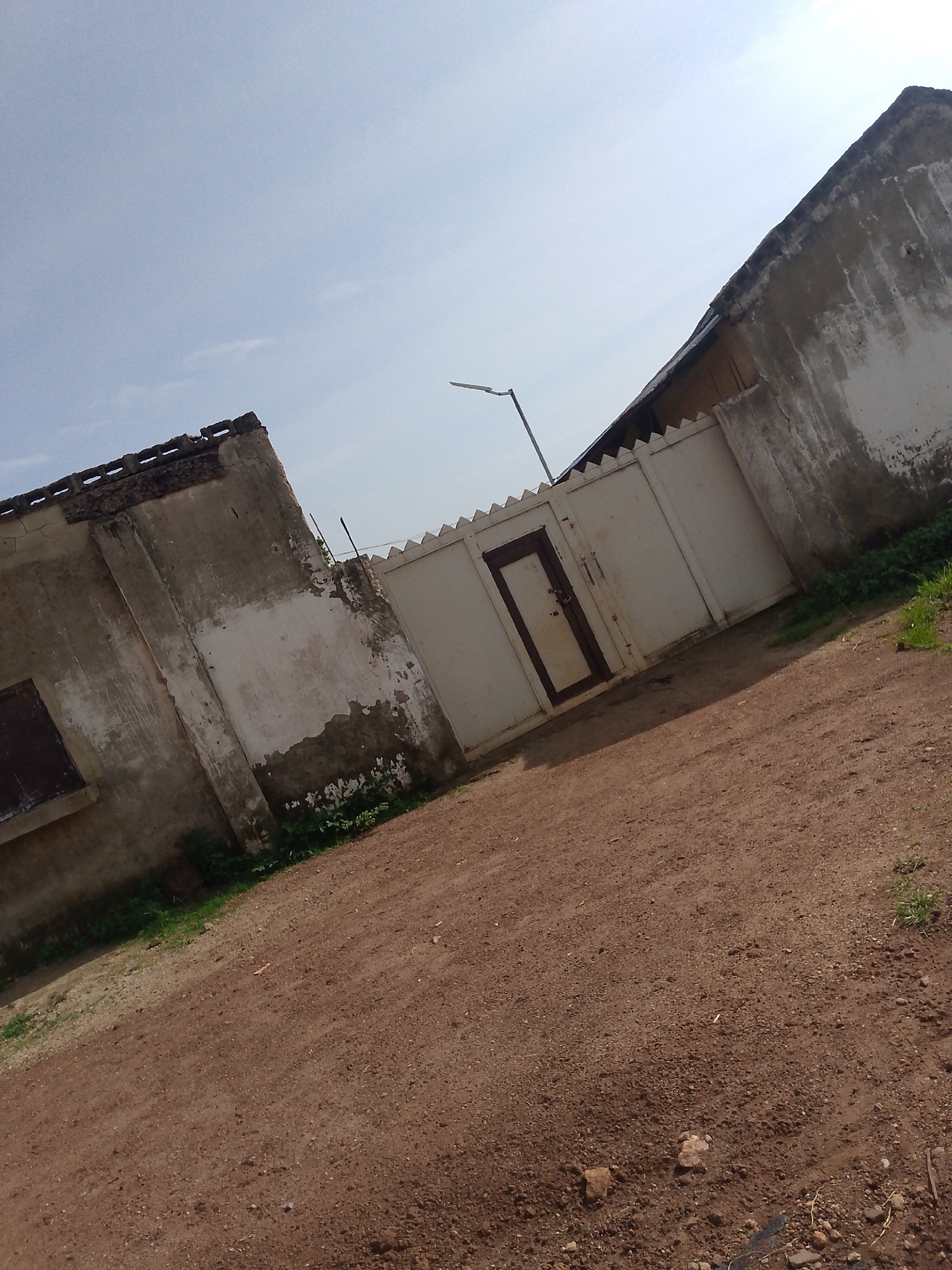 The image shows a property in Keffi, Nigeria. The foreground is a dirt road with some small stones and sparse vegetation. In the mid-ground, there is a white metal gate with a smaller door-like section on the left. The gate has a jagged top edge. Behind the gate and to the right is a wall, also with a jagged top edge. To the left of the gate, an old, weathered building is visible. This building has a dark-colored window and its walls are stained and peeling. The roof of this building is partially visible and appears damaged, with exposed bricks or blocks. Further to the right and behind the gate, another building or structure is visible, with a slanted roof. The sky is clear and light blue. A streetlight is visible on a pole in the upper mid-ground, to the left of the gate. The overall scene depicts an outdoor area with a gate, walls, and aging structures under a bright sky.