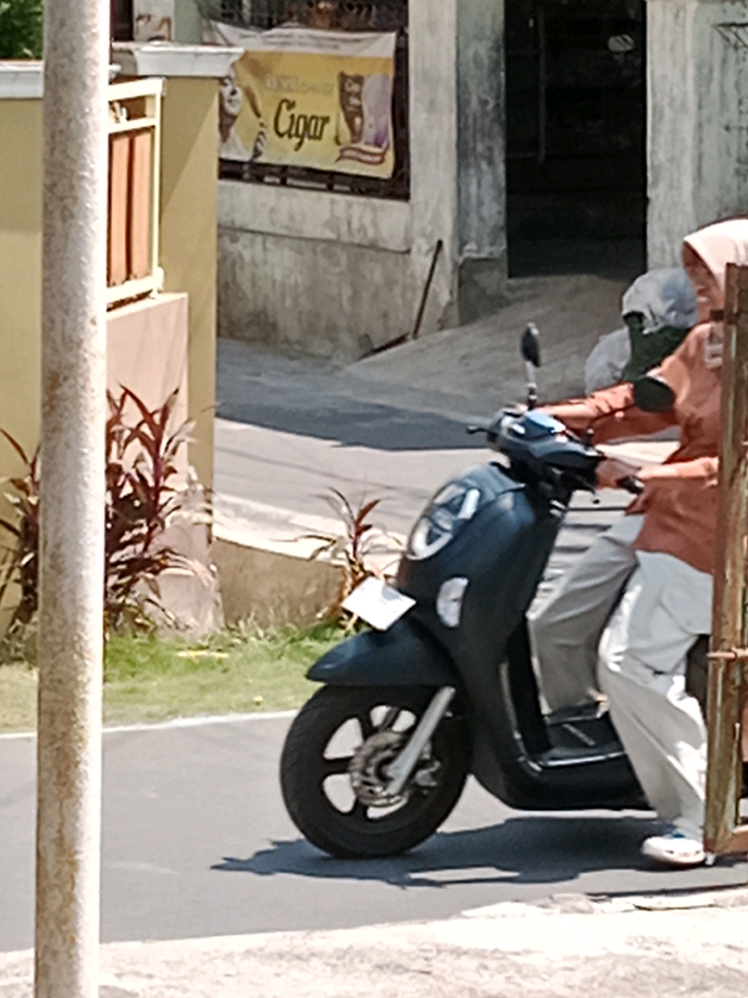 A medium shot captures a person riding a dark-colored scooter on a paved road. The rider, wearing a light brown hijab and an orange shirt, is partially visible from the chest down, with white pants and white shoes. They are holding the handlebars of the scooter. The scooter is positioned on the right side of the frame, with its front wheel and part of the body visible. To the left of the scooter, a tall, textured pillar stands prominently. Behind the pillar, a building with a yellow wall and a portion of a brown wooden structure is visible. In the background, a concrete wall with a banner advertising "Cigar" is displayed. The environment appears to be a street scene in Waru, Indonesia, with hints of a residential or commercial area. The road is paved, and there is some greenery and shadows on the ground.