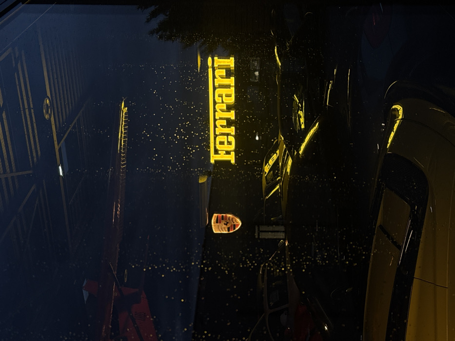 The image displays an exterior perspective, at night, looking into a car dealership showroom in Souffelweyersheim, France, through a glass window. The window surface is covered with numerous small water droplets, creating a diffused, bokeh effect across the view.
Inside the darkly lit showroom, two illuminated corporate signs are prominently featured. A horizontal, yellow neon sign spelling "Ferrari" in its characteristic font is positioned centrally. To its left and slightly above, an illuminated, shield-shaped emblem bearing the Porsche crest is visible.
Below these signs, multiple high-performance or luxury vehicles are parked. In the immediate foreground, the front section of a yellow sports car, including parts of its hood and fender, is visible, reflecting some light. Further within the showroom, additional vehicles, appearing as dark shapes with glints of reflected light on their surfaces, are indistinctly arranged; one appears to be a classic yellow sports car further back.
Reflections are present in the upper section of the glass, including a red angular object on the upper left and a horizontal structure with multiple segments above the Ferrari sign, possibly reflecting internal showroom architecture. Outside the showroom, a dark mass of foliage, likely a tree or dense shrubbery, is visible on the far right. No individuals are present in the scene.