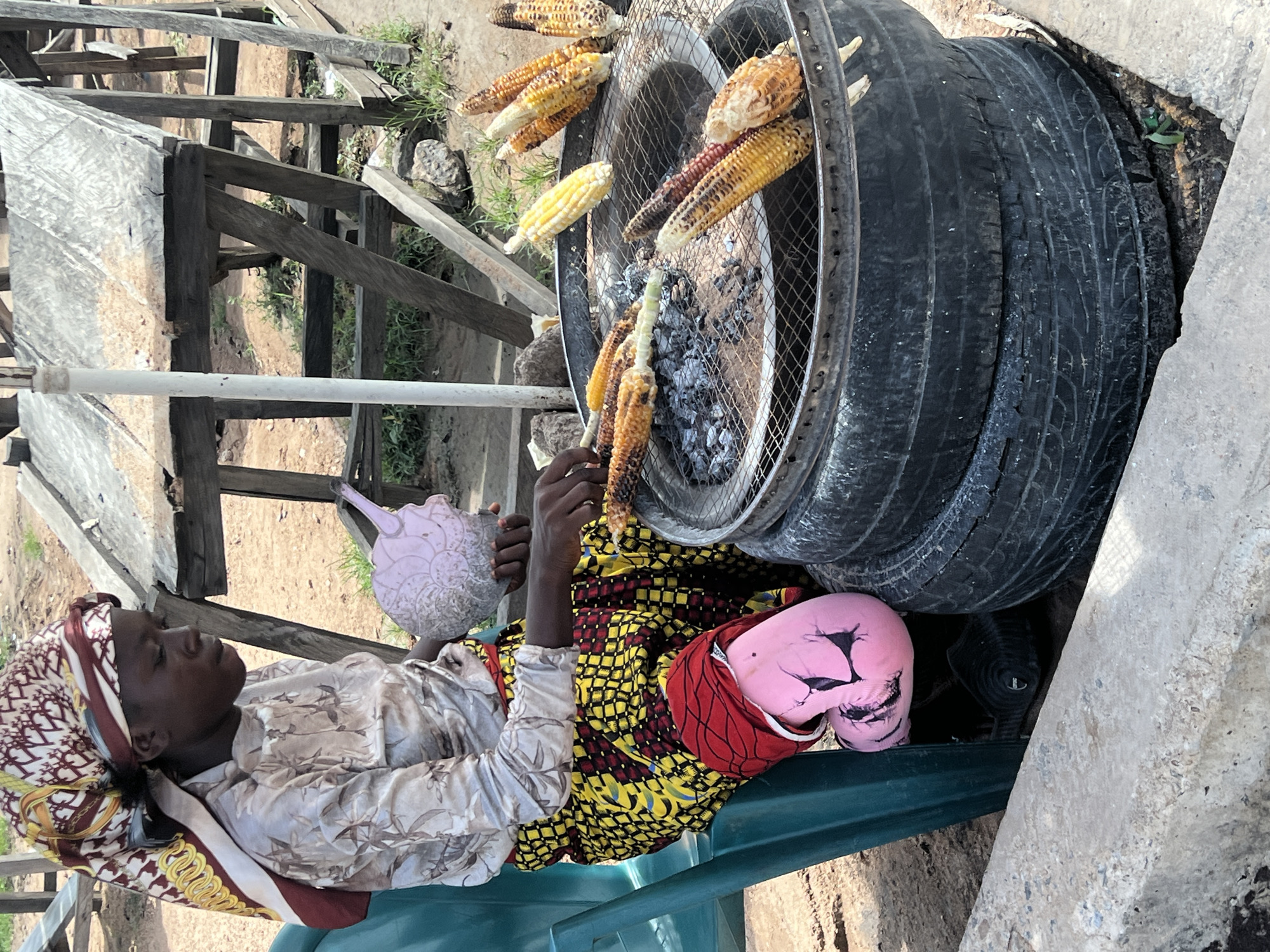 This detailed image captures an outdoor scene in Jalingo, Nigeria, depicting a woman engaged in grilling corn. A young Black woman sits on a teal plastic chair, positioned on the left side of the frame. She wears a multi-patterned headscarf featuring brown, white, red, and gold designs, a long-sleeved light beige shirt with a subtle floral or abstract pattern, and a vibrant skirt with a repeating pattern of yellow, black, and red squares. Her visible lower left leg is covered by a fitted pink fabric or garment with black markings. Her right hand holds a light purple, shell-shaped object, while her left hand manipulates a cob of corn over a grilling surface.
The grilling setup occupies the right side of the image. It consists of two stacked black vehicle tires forming a base, topped with a circular metal grate. Glowing coals and ash are visible beneath the grate. Multiple cobs of corn are being grilled on the grate, exhibiting varying degrees of charring; some are bright yellow, others show mixed yellow and reddish kernels, and several are significantly browned. Some grilled cobs are placed along the edge of the grate.
In the background, to the right of the woman and behind the grilling station, crude wooden structures with flat, weathered plank surfaces are visible, suggesting makeshift stalls or tables. The ground is dry, dusty earth with sparse green vegetation. A white vertical pole or pipe is partially visible on the left side, behind the woman. The scene is illuminated by bright daylight.