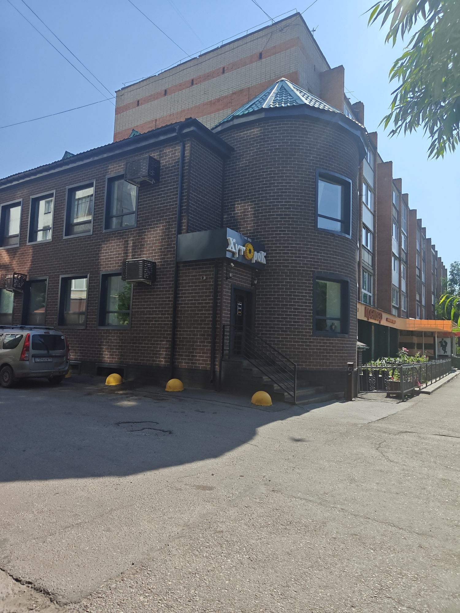 The image depicts the exterior of a building with dark brickwork, featuring several windows and air conditioning units. A silver minivan is parked in front of the building, and yellow spherical bollards are visible on the pavement. To the right, a section of the building has an orange awning extending over a sidewalk. In the background, a taller, multi-story building with beige and orange brick accents is partially visible. Green foliage from a tree extends into the upper right corner of the frame. The sky is clear and blue. A sign on the building reads "Хит Рок" (Hit Rock) with a sunflower logo. Another sign further down the street displays Cyrillic text, possibly indicating a business name. The scene is set on a paved surface, suggesting an urban environment.