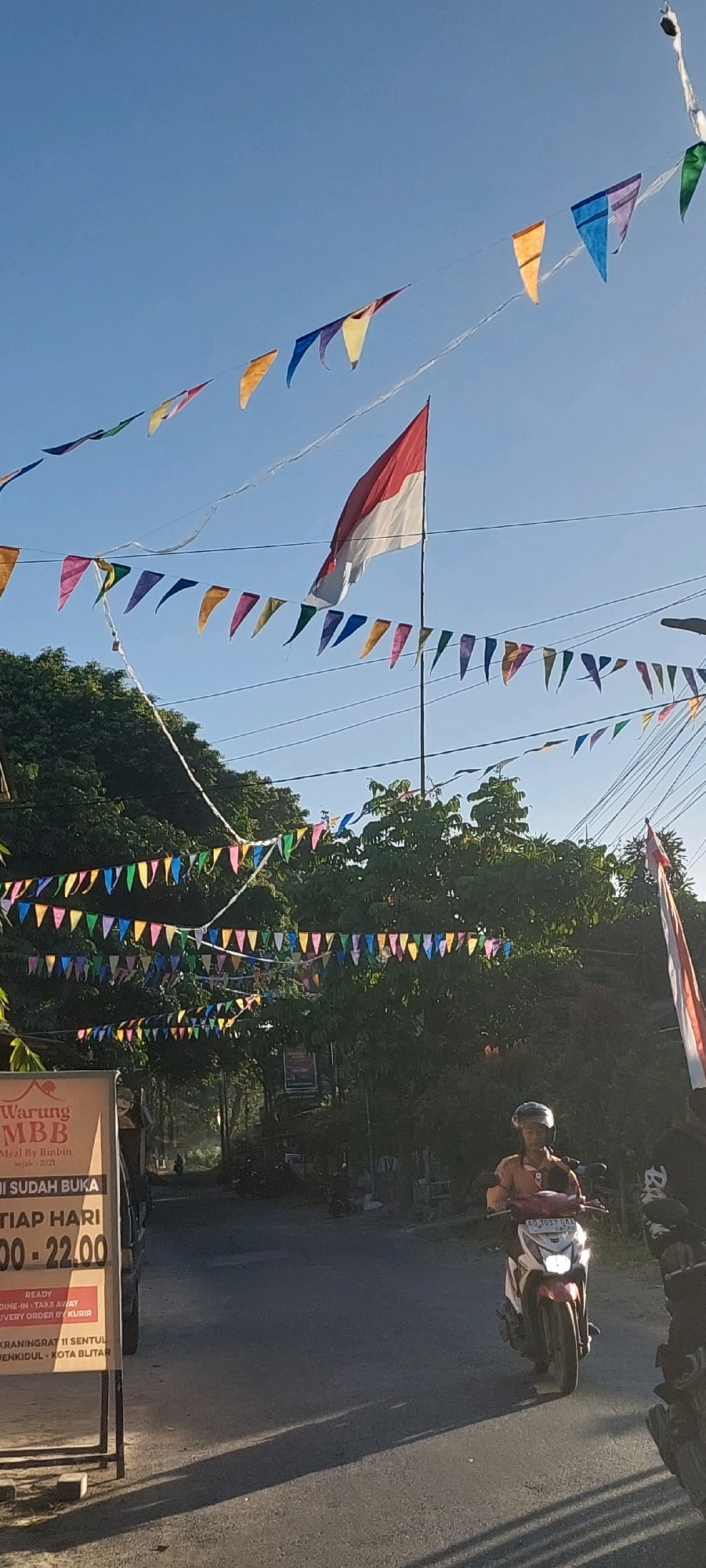 A vertical medium shot captures a street scene decorated with numerous colorful triangular flags strung across wires above a road. A large Indonesian flag, red and white, flies prominently from a pole in the center of the image, casting a shadow on the road. Several lines of small, multi-colored flags, predominantly in shades of yellow, blue, purple, and red, are also visible, creating a festive atmosphere. Lush green trees line the sides of the road, with one side featuring a prominent vertical sign for "Warung MBB" (Meal By Binbin, since 2021). The sign also indicates operating hours and delivery options. On the road, a person wearing a helmet rides a red and white scooter with its headlights on, moving towards the viewer. Another scooter is partially visible on the right side of the frame. The sky is clear and blue, indicating a sunny day. The overall scene suggests a local event or celebration in a street with overhead decorations.