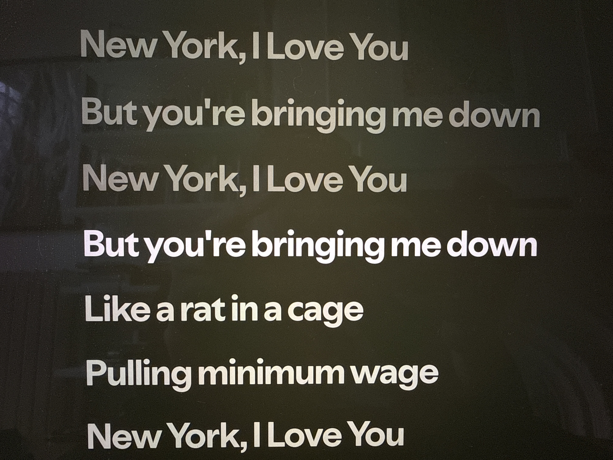 The image displays a digital screen in a New York City subway station, United States of America, showing white text against a dark grey or black background. The text consists of lines from the song "New York, I Love You But You're Bringing Me Down" by LCD Soundsystem. The visible lyrics are: "New York, I Love You", "But you're bringing me down", "New York, I Love You", "But you're bringing me down", "Like a rat in a cage", "Pulling minimum wage", and "New York, I Love You". The typography is a clean, sans-serif font. The screen surface shows faint reflections and subtle textural variations, particularly on the left side. No people, objects, or actions beyond the static text display are visible.