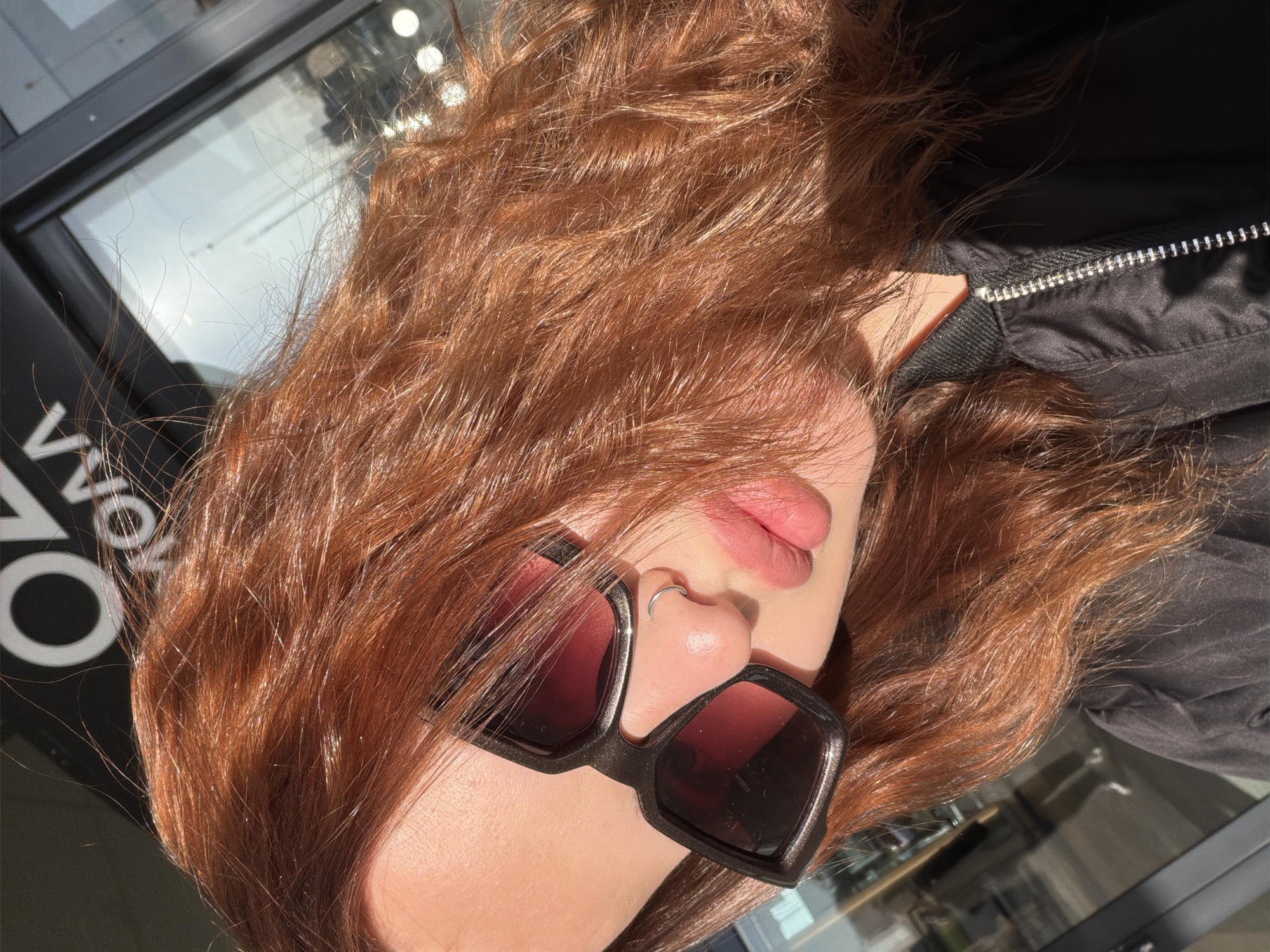 The image presents a close-up, angled portrait of a person, likely taken outdoors in bright sunlight. The subject's face is partially visible, dominated by long, wavy, reddish-brown hair that glows with golden highlights from the strong sun. They are wearing dark-framed sunglasses with reddish-tinted lenses, obscuring their eyes. A small, silver-colored hoop nose ring is visible on their left nostril, resting on the bridge of their nose. Their lips are slightly parted, showing a natural pinkish tone.
The person is dressed in a black jacket, parts of which are visible on the right and lower right of the frame, featuring a distinct silver zipper. The background appears to be a glass surface, reflecting bright light sources and indistinct shapes, possibly from an interior or opposing buildings. Partially visible white text, reversed due to reflection, can be seen on a dark background through the glass, resembling "VO" and fragments of other letters, suggesting signage. The bright, sunny weather is evident from the intense illumination and highlights. The scene is set in an urban or commercial environment in Krasnodar, Russia, likely during the daytime. The overall impression is casual and relaxed.