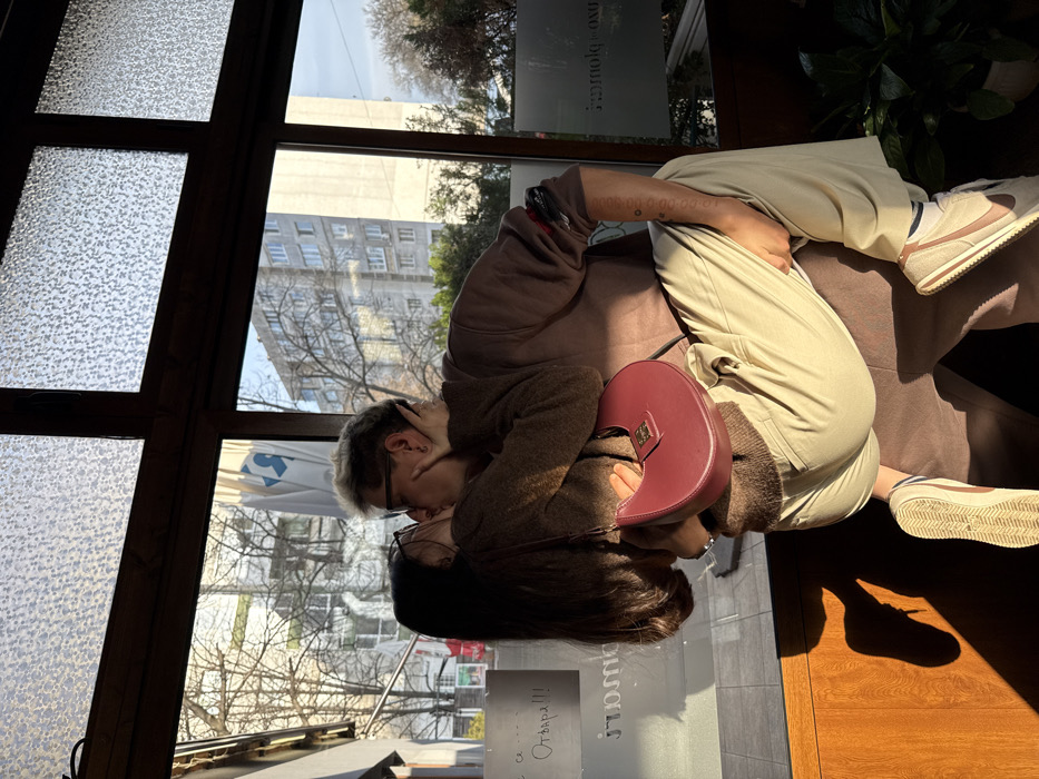 Two individuals are captured indoors through a large window, overlooking a city street scene. The location is identified as Sofia, Bulgaria, based on the reflected Bulgarian text "Отвара!!!" (Open!!!) and the characteristic multi-story urban architecture visible in the background.
In the foreground, a person with long dark hair, wearing glasses and a brown knitted sweater, holds a red crescent-shaped handbag with a gold-colored clasp. This individual is being lifted by another person with short bleached blonde hair, wearing glasses, a dark brown long-sleeved top, and light beige trousers. Both individuals are engaged in a kiss, with their hands positioned on each other's heads or necks. The individual on the right has visible tattoos on their left forearm. The individual on the left wears beige sneakers with white soles, while the individual on the right wears light-colored sneakers with a darker stripe.
The interior space features dark wooden window frames and a wooden sill. The upper sections of the windows are fitted with textured, frosted glass panes. Strong sunlight enters from the right, creating distinct shadows on the wooden sill and floor. Through the clear lower window panes, a street is visible with bare deciduous trees. Multiple multi-story residential or commercial buildings, typical of post-WWII Eastern European urban centers, are in the mid-ground. A large white flag or banner with a blue logo is reflected on the left side of the window glass. A potted green plant is visible in the lower right corner of the interior.
