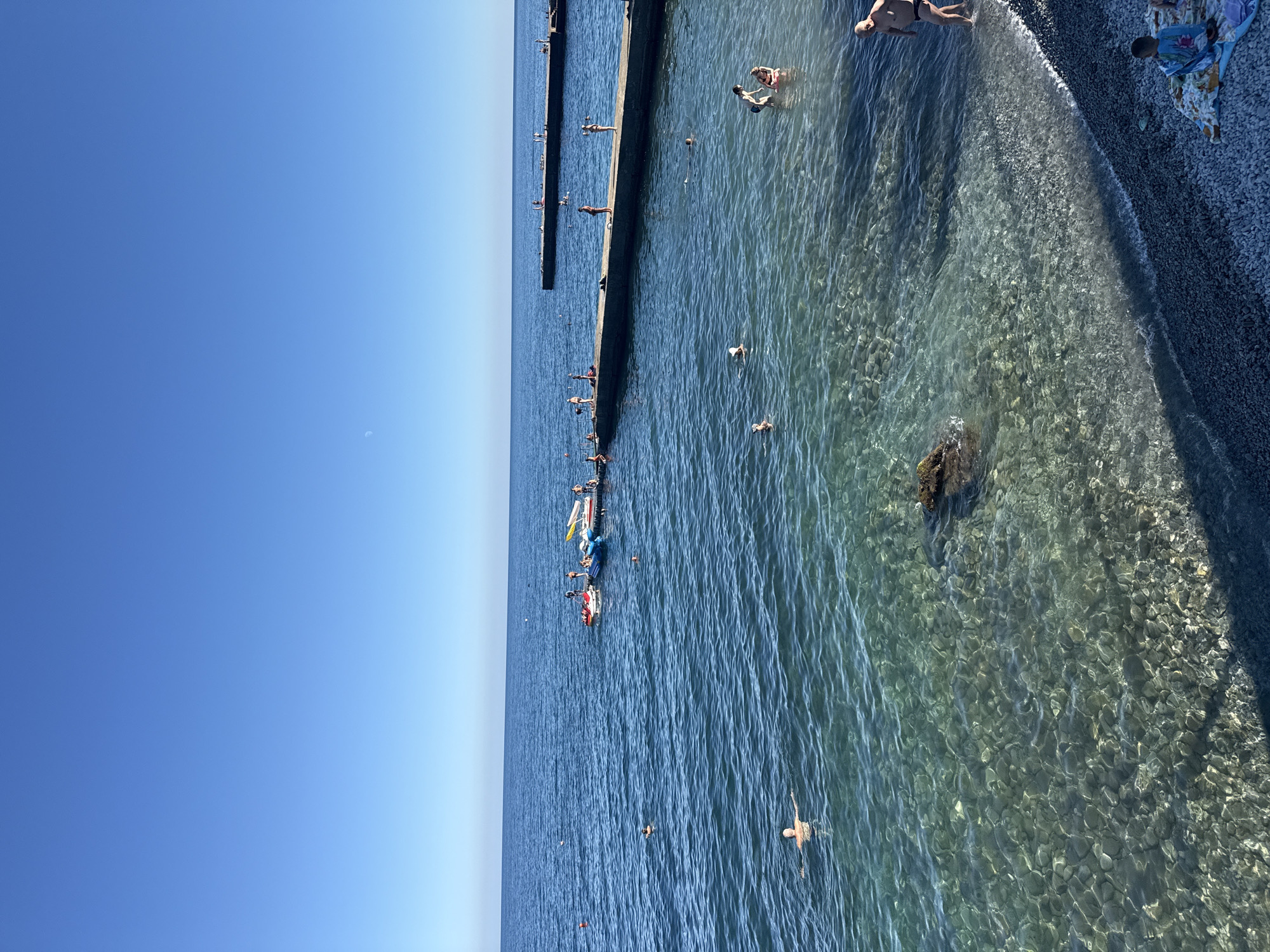 The image shows a view of the Black Sea coast in Partenit, Ukraine. The water is clear and blue, with visible pebbles on the seabed near the shore. Several people are enjoying the water and the beach. Some individuals are swimming in the sea, while others are standing or walking on the beach. There are also several jet skis and inflatable watercraft docked at a pier extending into the sea. The sky is clear and blue, with a faint crescent moon visible. Two concrete piers jut out into the water, with people on them as well. The beach itself is covered in dark grey pebbles.