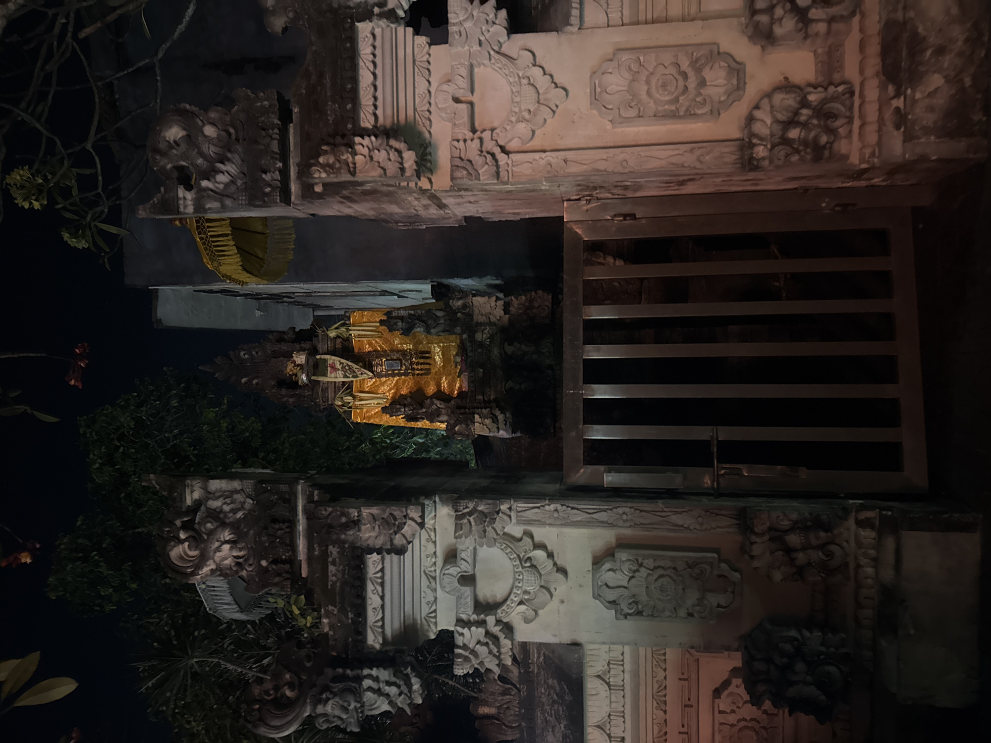 This is a nighttime, eye-level, medium shot of an ornate stone gateway with a metal gate, likely in Denpasar, Indonesia, given the architectural style. The gateway is flanked by intricately carved stone pillars, featuring decorative motifs such as floral patterns and monstrous faces. Beyond the gate, a structure is visible, adorned with a golden fabric and a statue or shrine. A yellow umbrella with fringe is positioned behind the shrine. The background is dark, with hints of foliage and a building. The overall impression is of a sacred or historical site at night, with atmospheric lighting enhancing the details of the stonework.