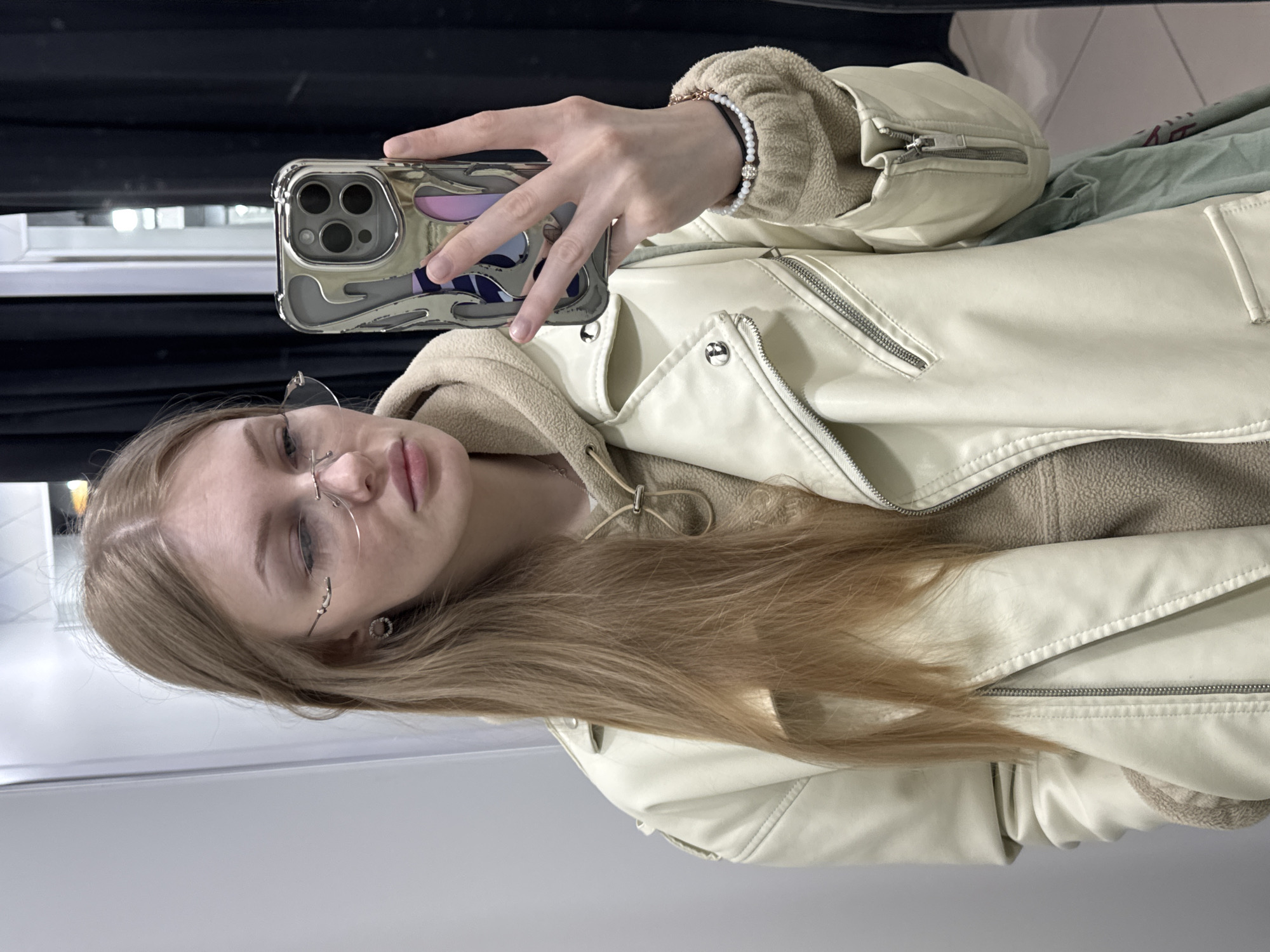 A young woman is captured in a mirror selfie, holding a silver-colored smartphone with a distinctive patterned case featuring visible camera lenses. Her head is tilted, and her long, light brown hair cascades over her shoulder. She is wearing rimless glasses and a neutral expression, possibly a slight pout.
She is dressed in a light cream or off-white faux leather jacket with multiple zippers and snap buttons, suggesting a biker or aviator style. Underneath the jacket, a beige or light brown hooded garment, possibly made of fleece or sherpa material, is visible. On her right wrist, she wears a bracelet composed of white beads and a darker strand, adorned with a small, decorative charm. A small stud or hoop earring is visible in her left ear.
The background appears to be an indoor setting, possibly a changing room or retail space, indicated by dark horizontal elements that resemble blinds or shelving, and plain walls or reflective surfaces from the mirror. The overall activity is a mirror selfie, with the woman extending her arm to hold the phone. No discernible text is visible in the image.
