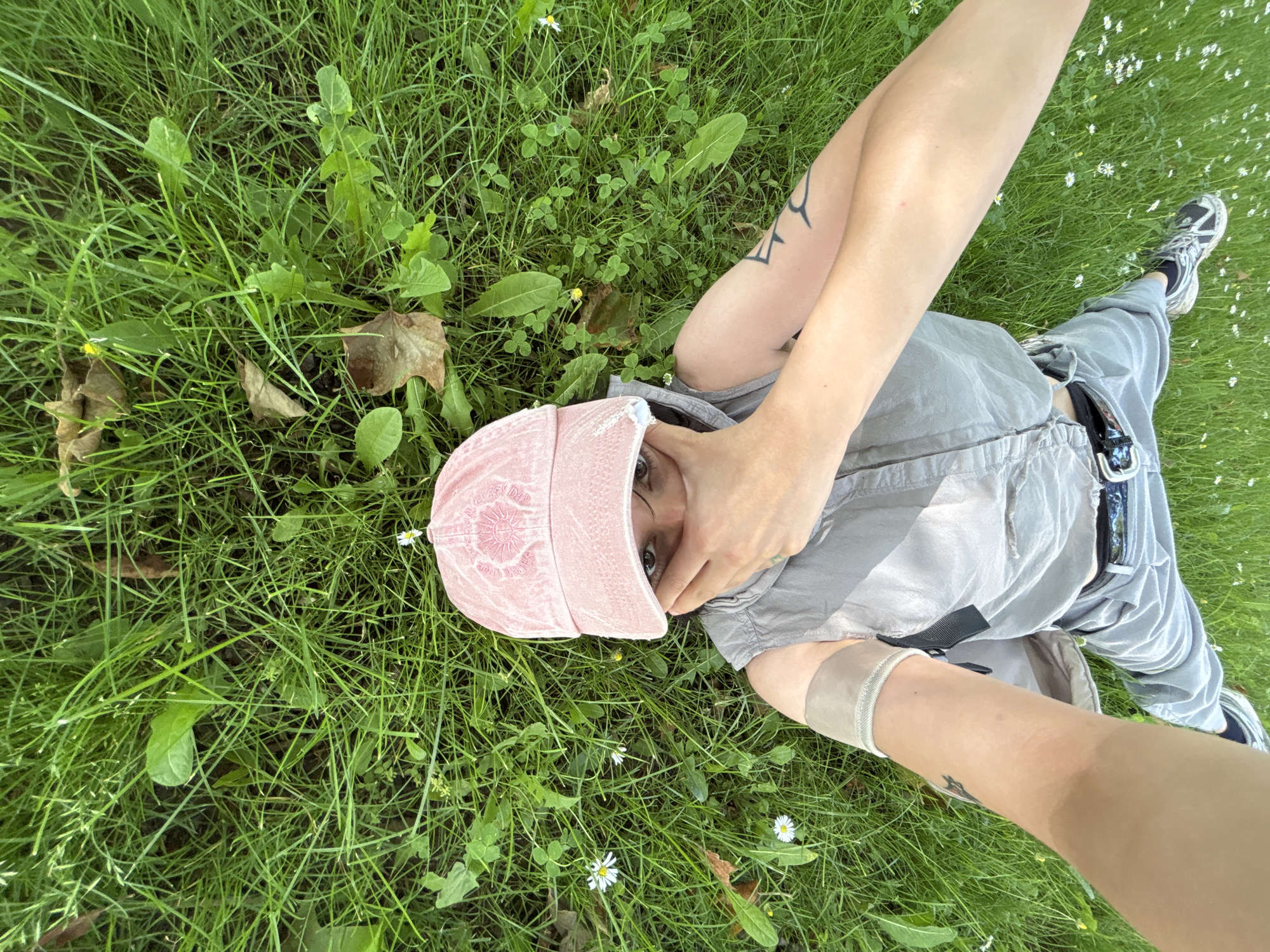 An individual lies supine on green grass in Sofia, Bulgaria, captured in an overhead wide-angle selfie. The subject, wearing a light pink baseball cap with a circular emblem, a gray sleeveless top, gray pants, and dark sneakers, holds the camera with their extended right arm. Their left hand covers the lower half of their face, exposing only their eyes beneath the cap's brim. A black tribal-style tattoo is visible on the left upper arm, and a light-colored band encircles the left forearm. The grassy environment contains various green plants, small white daisy-like flowers, sparse yellow blossoms, and scattered brown dried leaves.