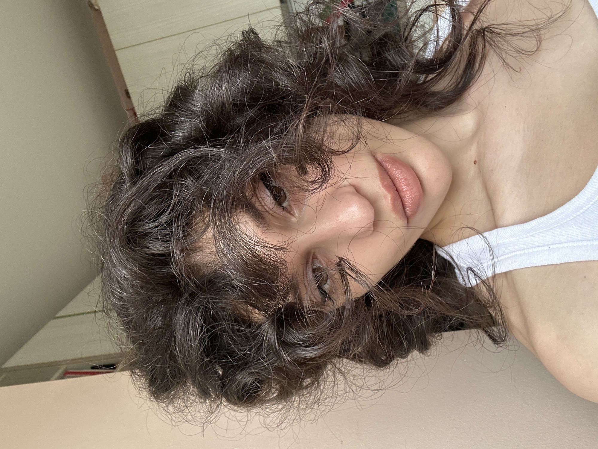 A young woman with dark, curly hair is pictured indoors, in what appears to be a residential setting in Sofia, Bulgaria. She is wearing a white tank top and her shoulders and upper chest are visible. Her face is partially obscured by her hair, but her lips, nose, and one eye are in focus. The background suggests an interior space with beige walls and cabinetry, possibly in a bedroom or living area. The lighting indicates it is daytime.