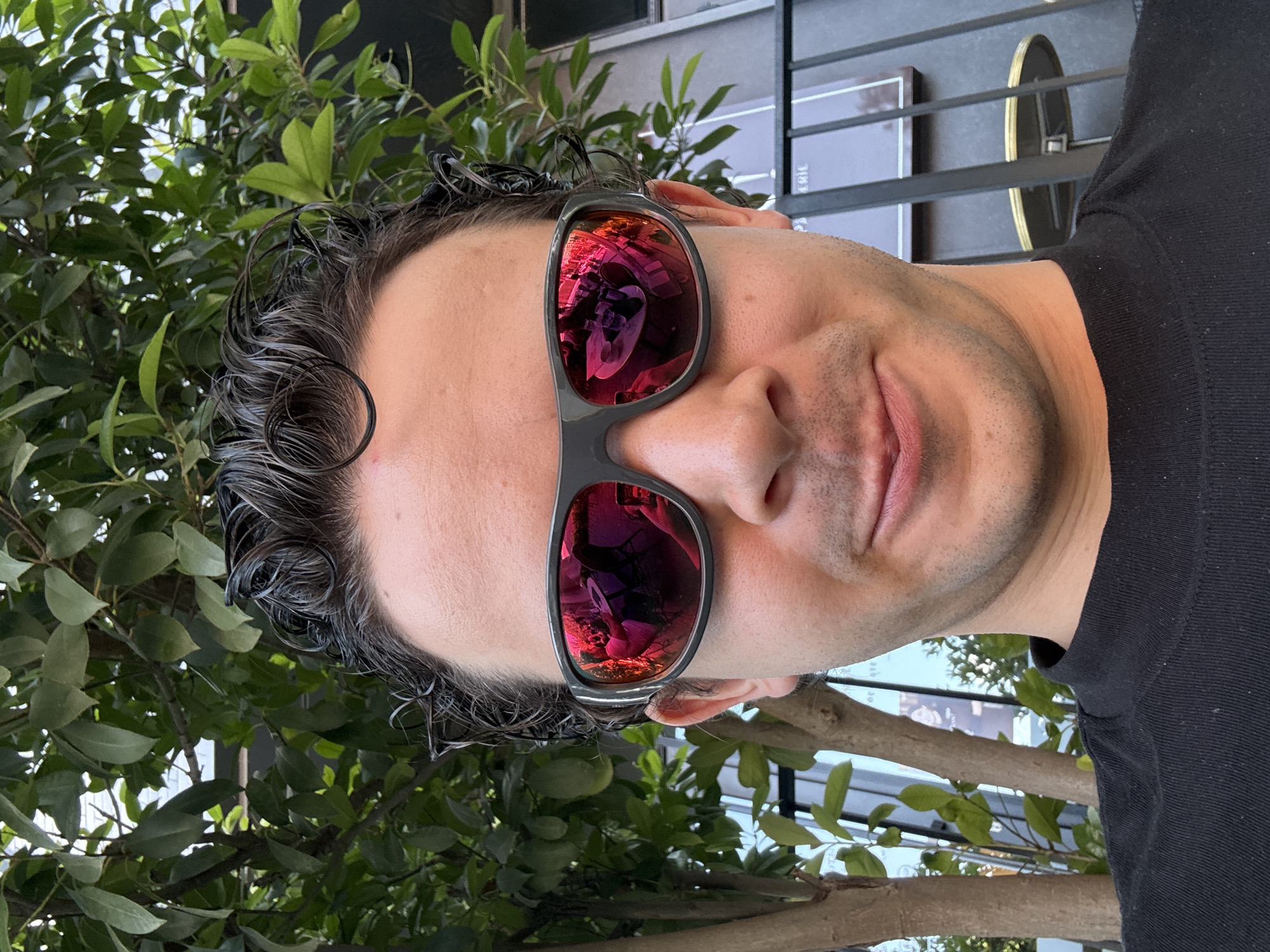 A man wearing sunglasses and a black shirt is posing for a selfie. The sunglasses have red-colored lenses and reflect an interior scene, possibly a restaurant. The man has short, dark, curly hair. In the background, there's a leafy green tree and a building with dark metallic bars and signage. The location is Tashkent, Uzbekistan, based on the reflection in the sunglasses.