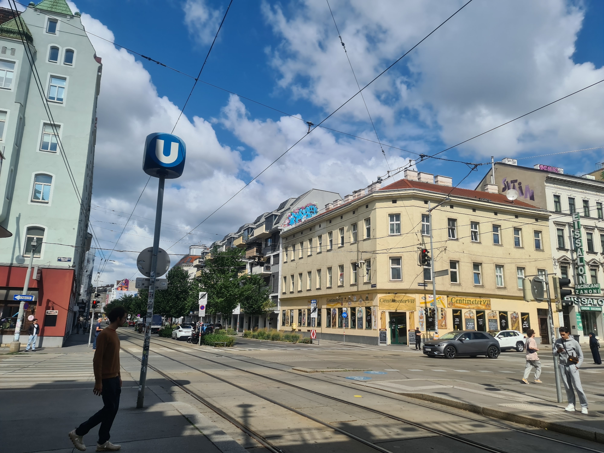 This is a street-level, medium shot photograph taken on a bright, partly cloudy day in Vienna, Austria. The scene captures a wide intersection with tram tracks running across it.
In the foreground, a young man wearing a brown sweater and dark pants is walking across the street from left to right. He is facing away from the camera, and his left leg is extended forward. To the right of him, a man in a grey tracksuit is standing on the sidewalk, looking down at his phone.
Several buildings line the street. On the left, a tall, light blue-green building with a green tiled roof occupies the corner. Across the street, a pale yellow building features multiple stories with numerous windows. The ground floor of this building houses shops, including one labeled "Centimeter VII." Graffiti in bright colors is visible on some of the buildings in the background.
A prominent blue U-shaped sign, indicating a subway station, stands on a pole near the center of the frame. Traffic lights are visible at the intersection, with one showing red. Several cars are present, including a dark grey SUV and a white SUV. A few other pedestrians are scattered throughout the scene, and a motorbike is parked on the right side of the street. Overhead, tram wires crisscross the sky, and trees with green foliage are visible along the sidewalks. The overall impression is a typical urban street scene in a European city.
