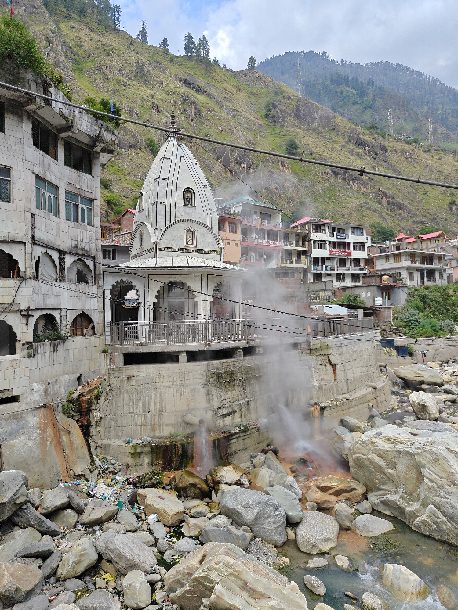 This media file presents a bustling scene in a mountainous region, likely Kasol, India, featuring buildings clustered along a steep hillside beside a river. In the center, a prominent white temple with a conical spire (shikhara) and arched entrances stands on a concrete foundation. Adjacent to it on the left is a multi-story white building with several windows and arched openings on its lower levels.
In the foreground, a rocky riverbed with a flowing stream is visible. Numerous plumes of steam rise from hot springs emerging from the base of a concrete retaining wall, staining the surrounding rocks a reddish-orange hue. Unfortunately, a considerable amount of litter, including plastic bottles and bags, is scattered among the rocks in the left portion of the riverbed. Utility lines stretch across the mid-ground.
The background reveals a steep, green, and rocky mountainside, dotted with trees, extending upwards towards an overcast sky. More buildings, seemingly guesthouses or residences with balconies and varied roof colors (some red), are nestled into the hillside behind the main structures. Text visible on some of these buildings includes "PADMA VILLA GUEST HOUSE" with "PANKAJ RESTAURANT" below it, and partially, "HO DAY GUEST HOUSE". The scene is captured during daylight hours under cloudy weather conditions. No people are discernible in the image.