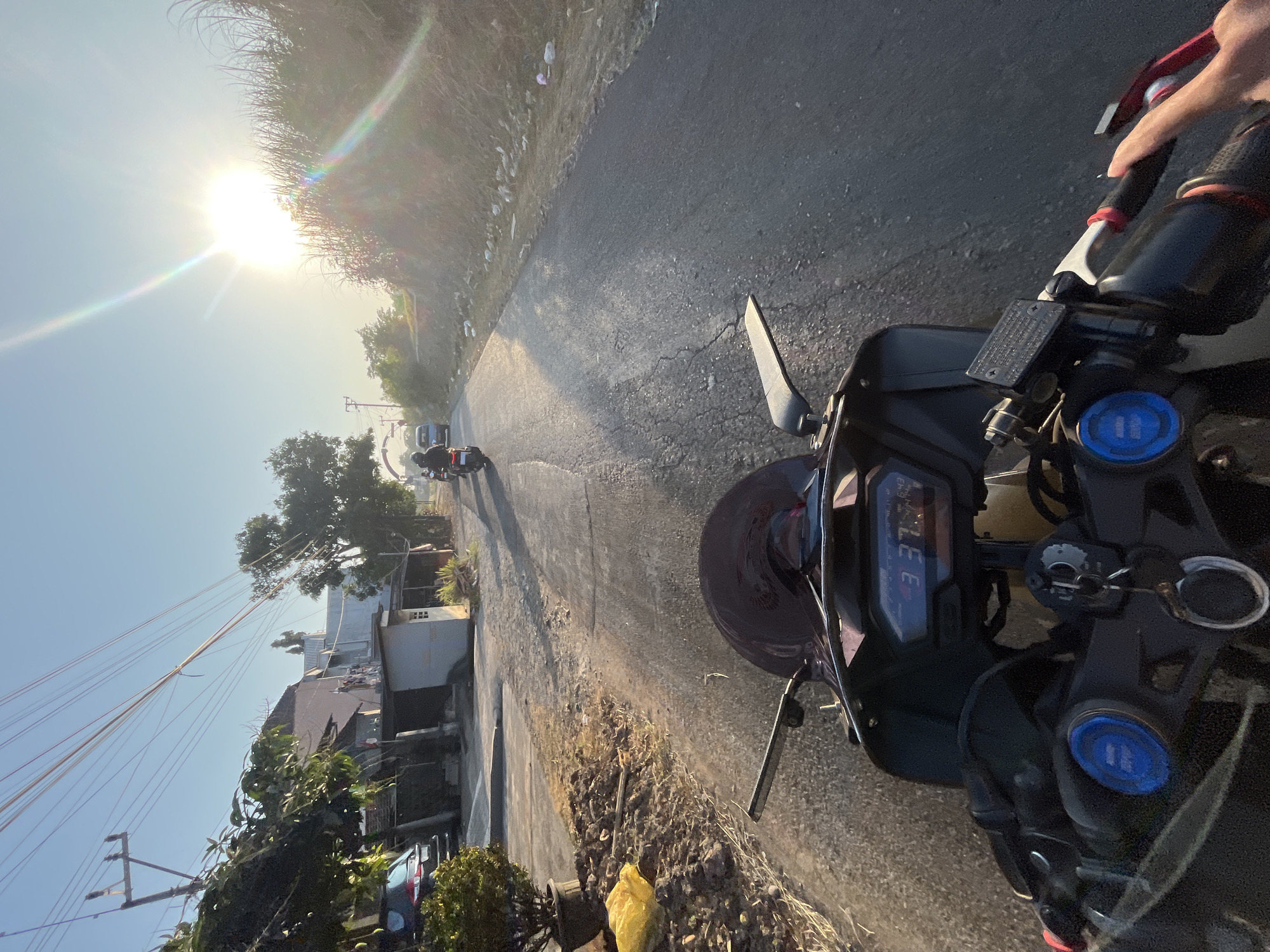 The image depicts a first-person perspective of someone riding a motorcycle on a paved road in Pecangaan, Indonesia, during what appears to be early morning or late afternoon due to the low angle of the sun and the long shadows. The road is cracked and shows signs of wear. To the left of the road, there are houses, trees, a parked car, and a potted plant with a yellow plastic bag beside it. To the right, there is a grassy embankment with some scattered debris. Other motorcycles are visible further down the road. The motorcycle's digital dashboard shows the time as 3:37. The sky is clear and bright, with the sun creating lens flares. The overall scene suggests a rural or semi-urban setting.