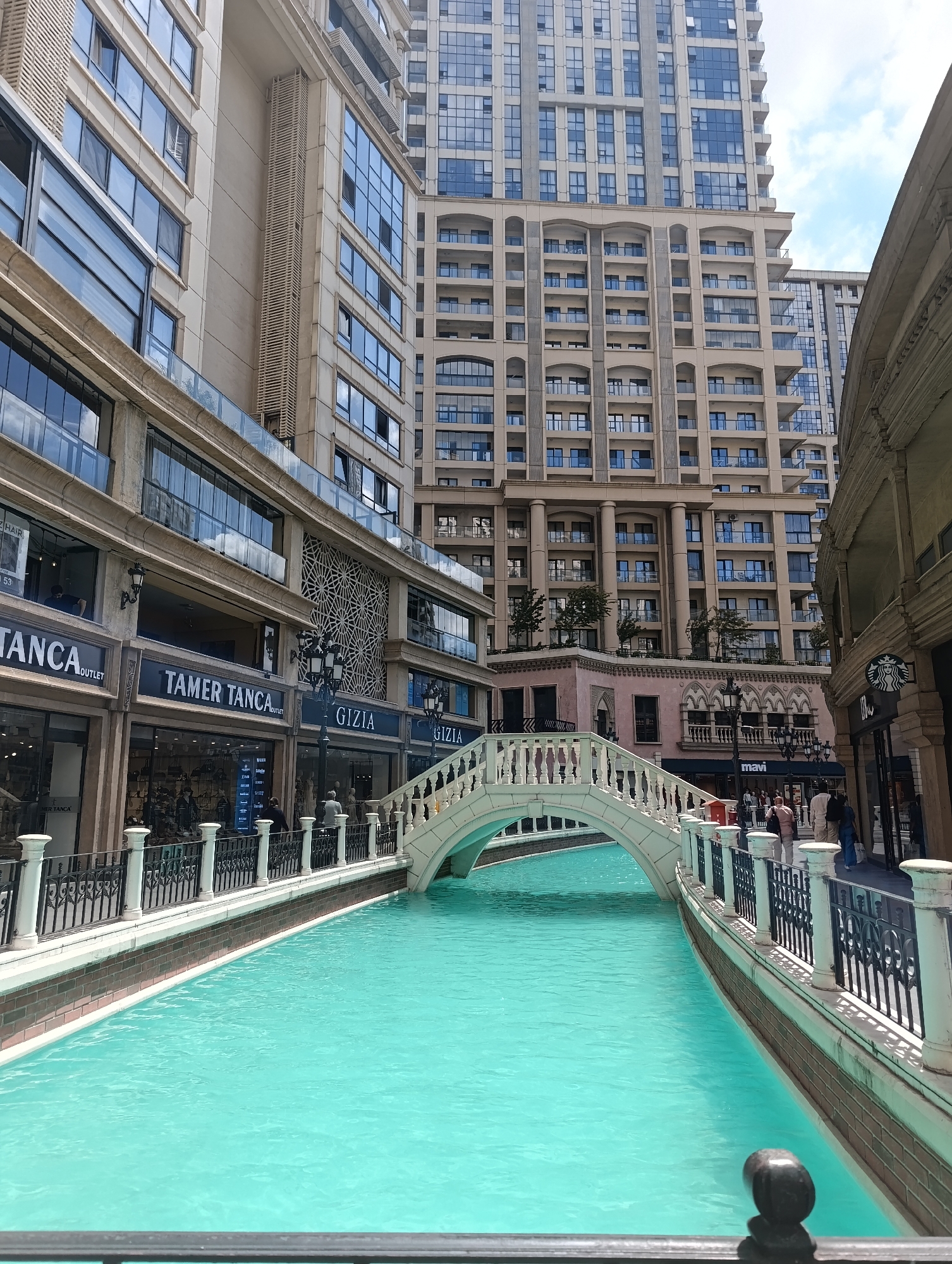 The image shows a brightly lit outdoor scene with a canal-like body of water in the foreground. The water is a vibrant turquoise color and appears calm. A white arched bridge spans the water, with ornate railings on either side. Along the banks of the water are walkways and storefronts. The buildings are tall and modern, with many windows. Signs for businesses such as "TANC A OUTLET," "TAMER TANCA," and "GIZIA" are visible on the storefronts. In the background, more modern buildings rise into the sky under a partly cloudy sky. Several people are walking along the walkways, dressed in casual clothing. The overall impression is that of a commercial or entertainment district designed to evoke a Venice-like atmosphere. This scene is located at the Venezia Mega Outlet in Gaziosmanpaşa, Istanbul, Turkey.