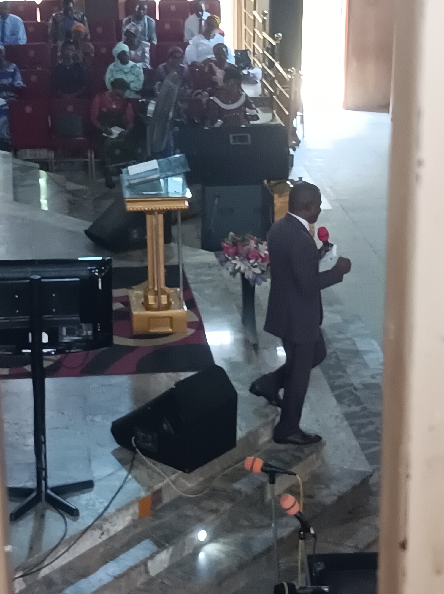 The image shows a man in a suit speaking into a red microphone inside what appears to be a church or religious building. The man is walking on a slightly raised platform. Seated people are visible in the background, facing the direction of the speaker. There is a lectern with books on it, speakers on the floor, a television set, and some floral arrangements nearby. The location is possibly in Dadi Riba, Nigeria.