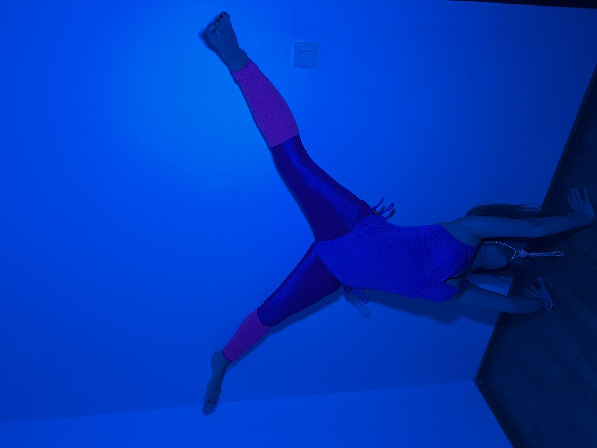 A woman in a blue leotard and shiny pink leggings is performing a split against a wall in a room lit by blue light. One leg is extended upwards, close to a wall switch, while the other leg is extended downwards. She is leaning forward, her hands touching the wall. A string of pearls adorns her neck. The location is presumed to be in a room in Budva, Montenegro, based on the user's prompt, although there are no identifiable landmarks or visual cues to confirm this definitively.
