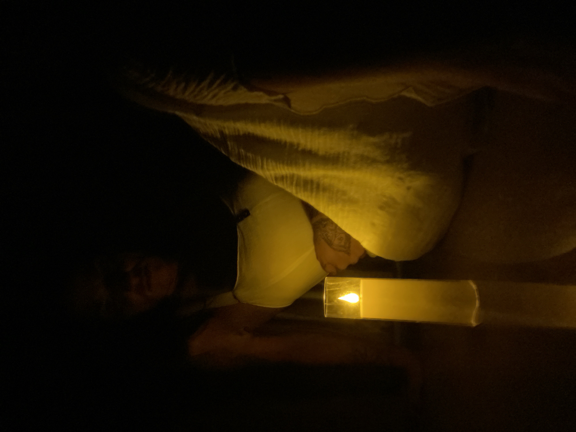 In the dimly lit environment of a room in Mũi Né, Vietnam, a solitary, tall, cylindrical candle with a lit wick glows warmly, casting a yellow luminescence. The candle stands in a clear glass holder on a reflective surface. To the right of the candle, a person is partially visible, reclined and draped in a flowing white garment that exhibits textured folds. The person's upper body is supported by a white undergarment. A tattoo is discernible on the person's left forearm. The background is almost entirely obscured by darkness, with only faint outlines of the person's facial features visible in the upper portion of the image. The overall scene is characterized by low light and the prominent presence of the illuminated candle.