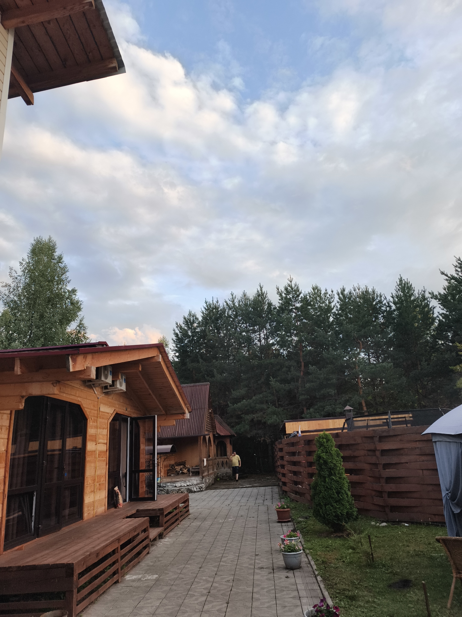 The image depicts a view of a resort complex in Katun, Russia, likely during the daytime or early evening. The scene is dominated by wooden structures, including a prominent, multi-story building with a wooden facade and a dark-shingled roof in the foreground, featuring large windows and an attached wooden deck. Further back, another smaller wooden building with a similar architectural style is visible, along with a pathway made of paved stones.
A lone individual, dressed in a yellow shirt and dark pants, is walking away from the camera on the pathway, towards the trees. The background is characterized by a dense line of coniferous trees, creating a natural backdrop. A wooden fence, constructed from horizontal planks, runs along the right side of the image, bordering a grassy area with potted plants and a small evergreen tree. In the far right, a portion of a gray canopy or gazebo is visible. The sky above is partly cloudy, with a mix of white and gray clouds against a blue sky. The overall impression is of a rustic, nature-oriented retreat.