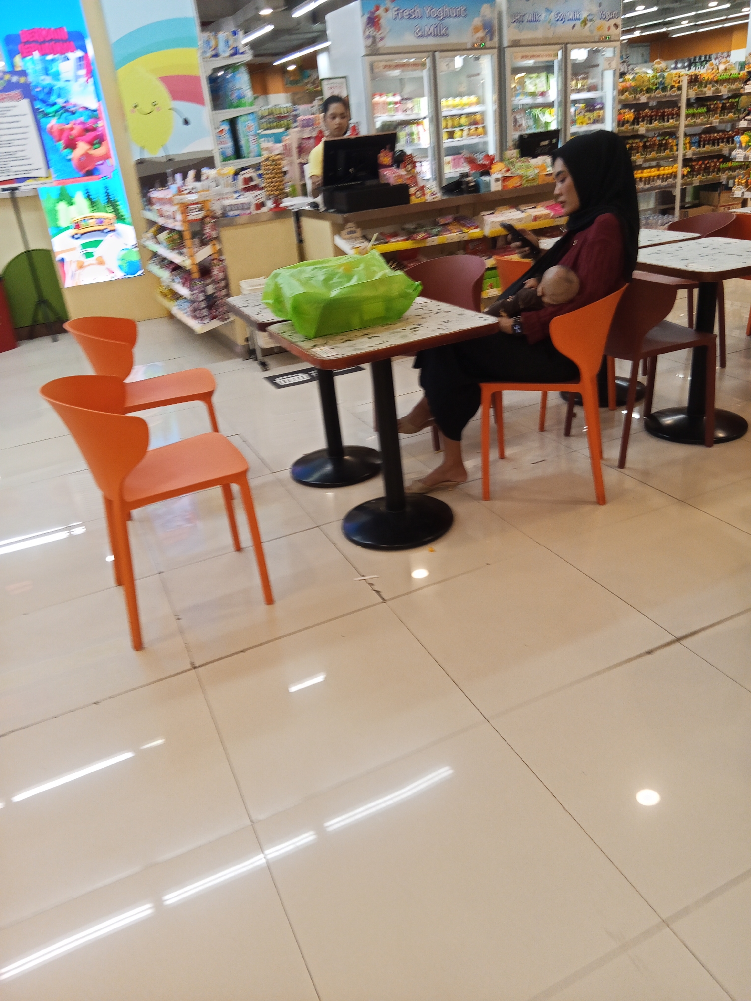The image depicts an indoor scene, likely a supermarket or a food court area, with a tiled floor reflecting overhead lighting. In the foreground, there are several orange plastic chairs around small, round tables. One table has a green plastic bag on it. In the mid-ground, a woman wearing a black hijab and a maroon top is seated at a table, holding a baby. She is looking down at her phone. To the left, a person in a yellow shirt stands behind a counter. Shelves stocked with various products are visible in the background, including refrigerated units displaying dairy products. The overall environment appears to be a commercial establishment. The location is identified as Lubuklinggau, Indonesia.