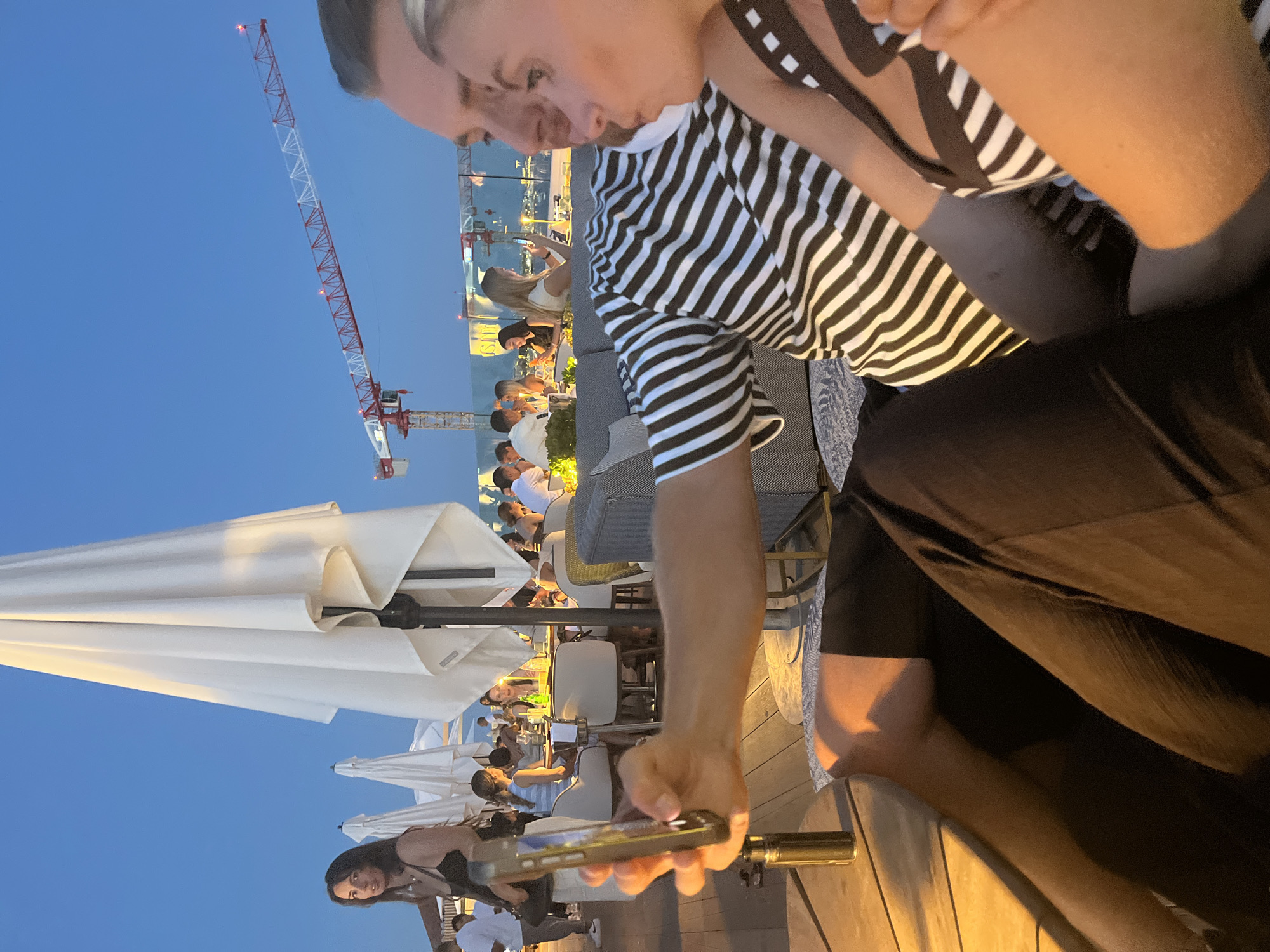The image depicts a rooftop restaurant or bar in Cannes, France, at dusk. In the foreground, a couple is seated, with the man holding a smartphone to take a selfie with the woman. They are dressed in black and white striped clothing. Behind them, other patrons are seated at tables, and white umbrellas are open. In the background, a construction crane is visible against the blue sky, suggesting ongoing development in the city. The overall atmosphere is relaxed and social, characteristic of a seaside dining establishment.