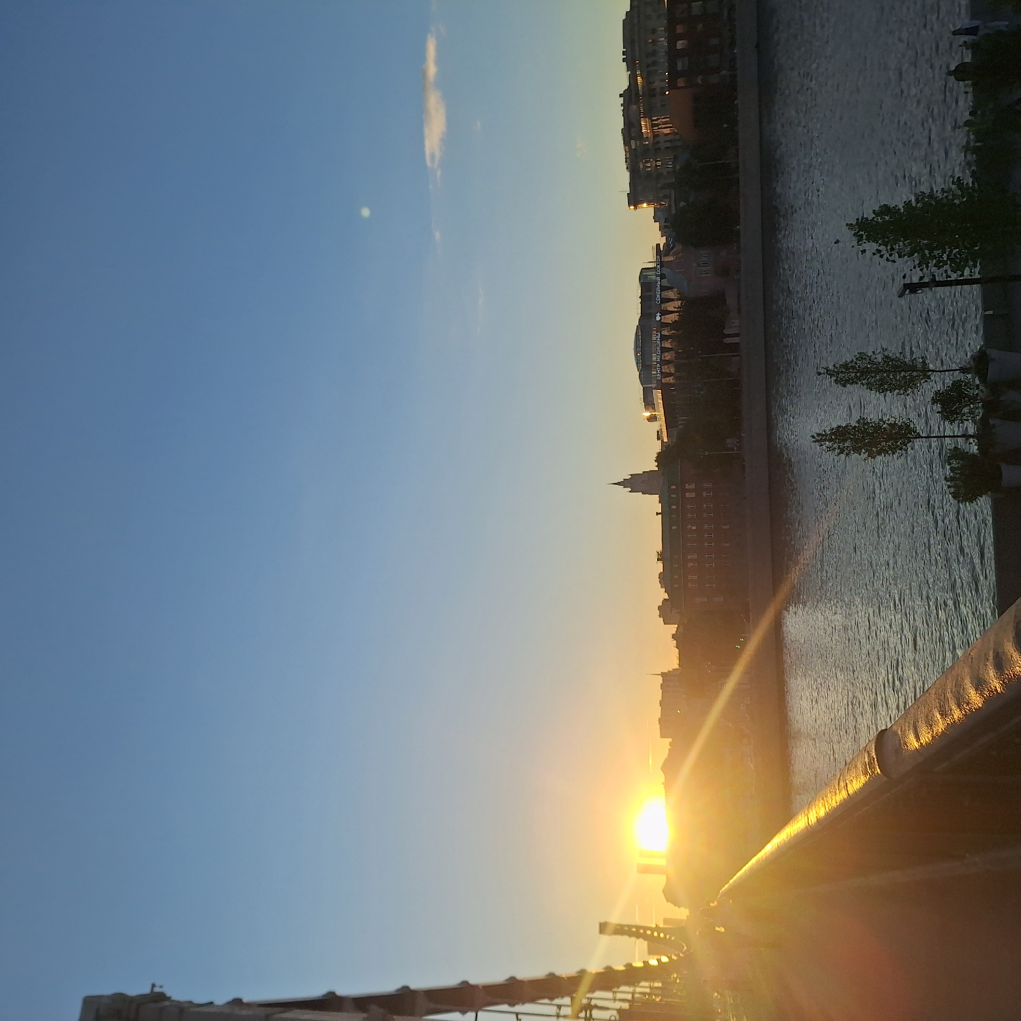 The image depicts a sunset view from a bridge in Moscow, Russia. The sun is setting over the city skyline, casting a golden glow on the buildings, water, and bridge railings. The Moscow River runs along the right side of the image, with buildings lining its bank. The bridge railing is visible in the foreground. The sky is clear blue, transitioning to a warmer hue near the horizon where the sun is setting.