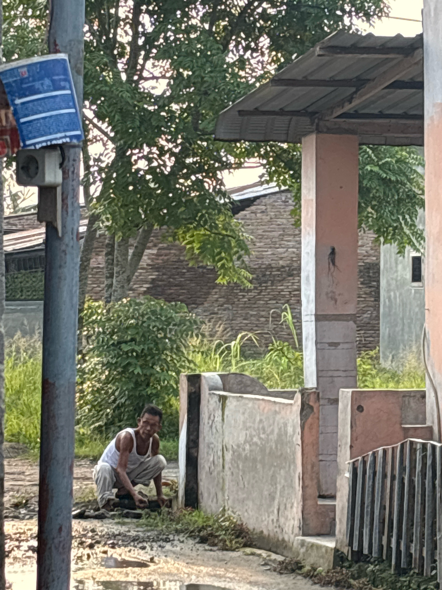 A man wearing a white tank top and light-colored pants is crouching on the ground in what appears to be an outdoor, possibly unpaved area. He is positioned near a low, concrete wall or barrier. The ground around him is wet and uneven, with puddles reflecting the surrounding environment. He appears to be interacting with something on the ground in front of him, possibly items laid out on the dirt. In the background, there are trees, a brick building, and part of a structure with a corrugated metal roof supported by a pillar. A utility pole with a blue and white container attached is visible in the foreground to the left. The overall setting suggests a semi-urban or rural environment, potentially within Helvetia, Indonesia, given the architectural styles and vegetation.