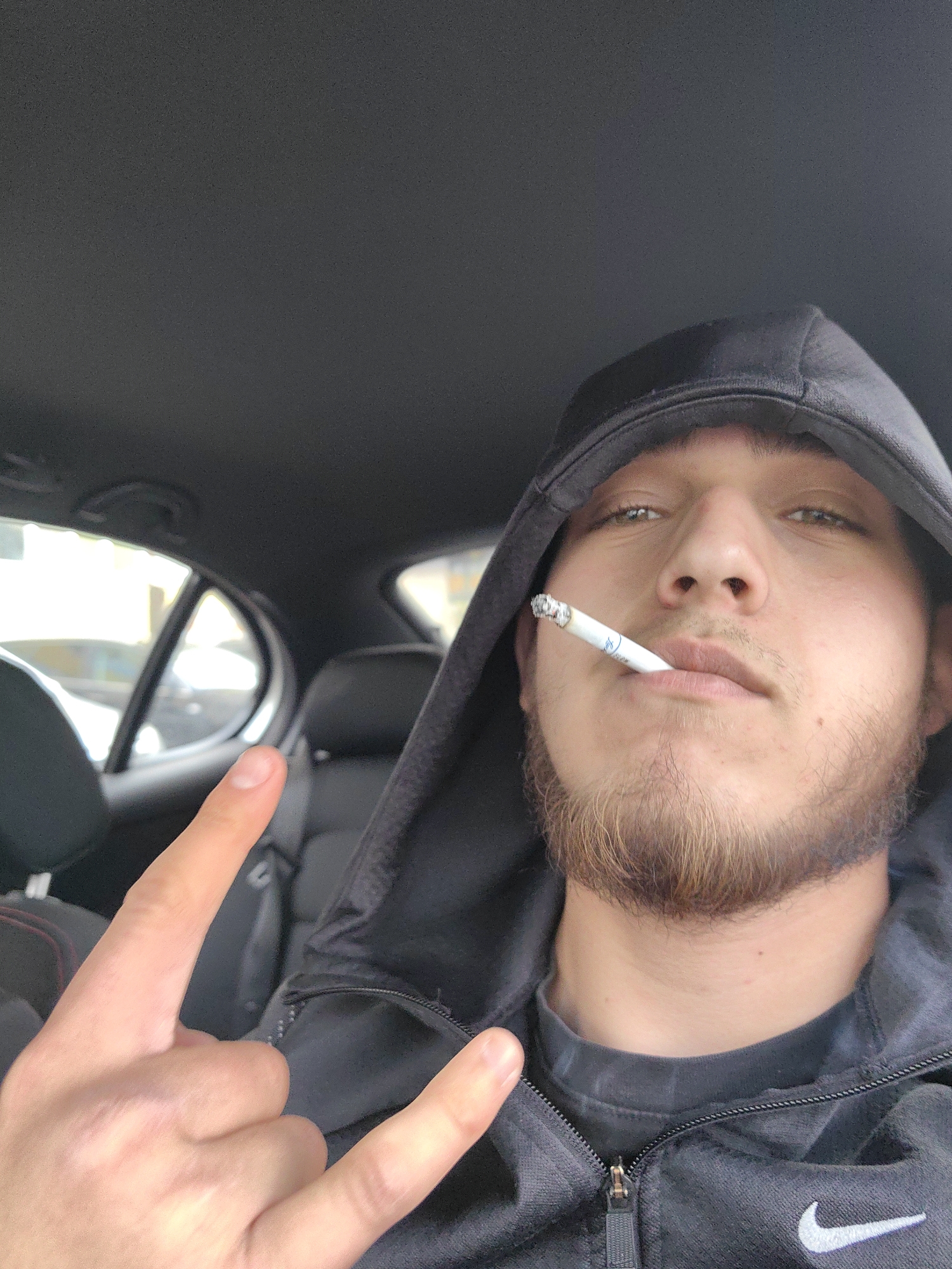 A young man is taking a selfie indoors, likely in a car. He is wearing a dark hoodie with the hood up and a gray t-shirt underneath. He has a beard and is holding a lit cigarette in his mouth. He is making a hand gesture with his left hand, holding up his index and pinky fingers, while the middle and ring fingers are curled. The background shows the interior of a car, including seats and windows. The location is likely not Sofia, Bulgaria as there is no information indicating it.