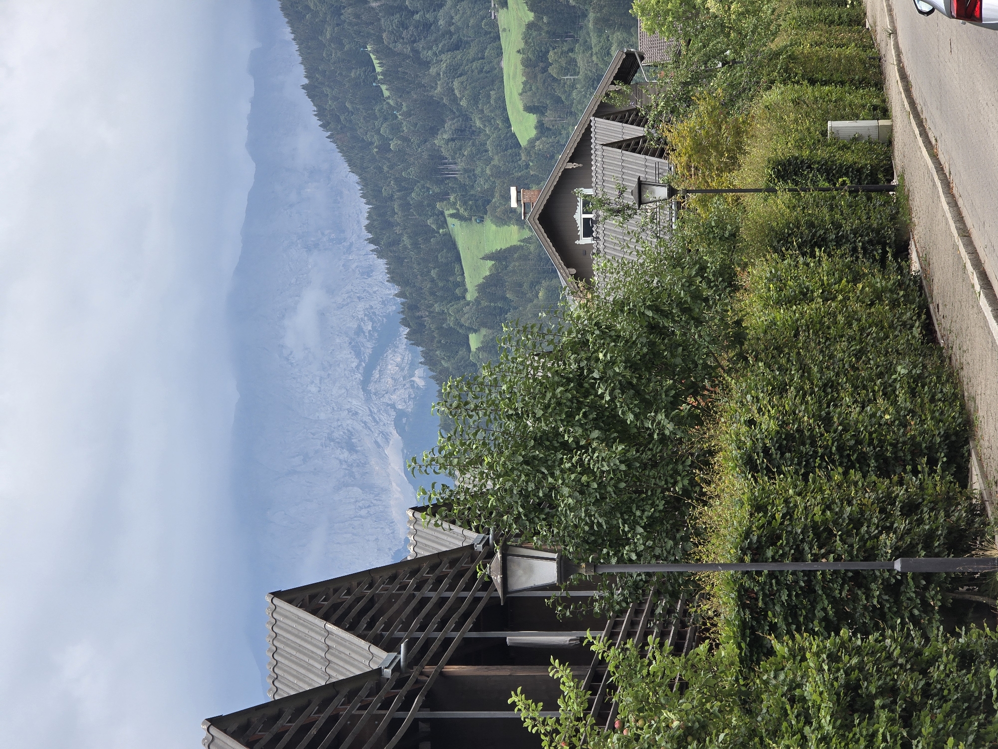 The image depicts a street scene in Garmisch-Partenkirchen, Germany, with a row of residential buildings and lush greenery. In the foreground, a paved sidewalk runs alongside a street, with a neatly trimmed hedge bordering the sidewalk. A dark lamppost stands tall beside the hedge, casting a subtle presence. Behind the hedge, the roofs of several buildings are visible, characterized by their dark, tiled surfaces. Some of these buildings feature wooden balconies or verandas, adding a rustic charm to the architecture. Trees and bushes, with their dense foliage, create a natural barrier between the street and the houses. In the background, a verdant hillside is covered with a dense forest, ascending towards the majestic, mist-shrouded mountains. The sky is overcast, lending a soft, diffused light to the scene. The overall impression is one of a tranquil, well-maintained residential area nestled amidst a picturesque natural landscape.