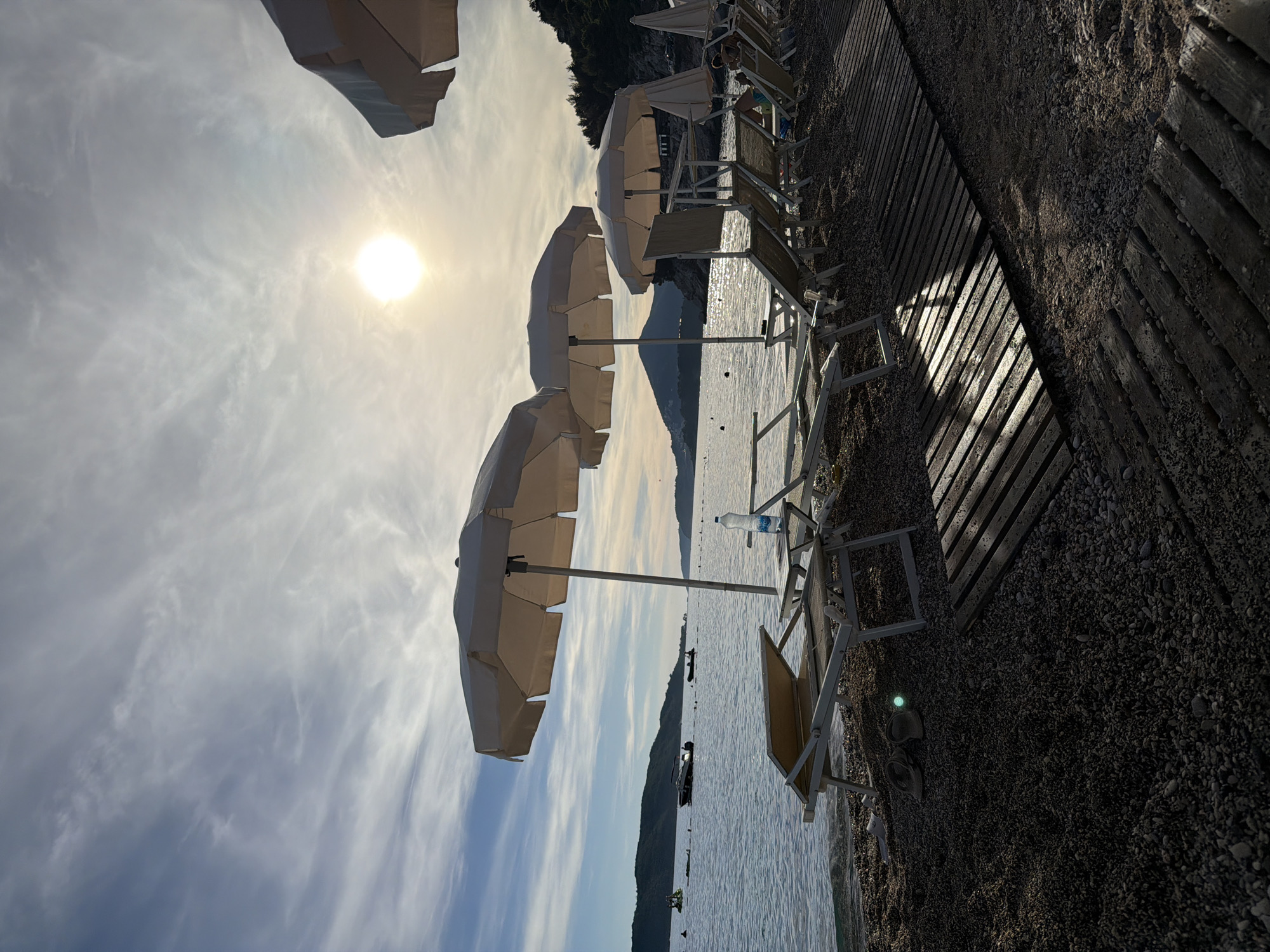 This image captures a serene beach scene in Kamenovo, Montenegro, during what appears to be late afternoon or early evening, indicated by the low angle of the sun. The foreground is dominated by a pebble beach, with wooden boardwalks laid out to provide access across the stones. Several empty sun loungers, some with white frames and beige fabric, are arranged on the beach, some positioned under large, cream-colored umbrellas.
In the middle ground, calm, blue water stretches towards a distant coastline lined with green, tree-covered hills. Several boats are visible on the water, including a white motorboat and a smaller dinghy. A few people can be seen in the water, and one person is seated on a lounger further down the beach. The sun, a bright orb, is visible in the upper right portion of the sky, partially obscured by wispy white clouds. The overall atmosphere is tranquil and inviting, showcasing a typical European coastal resort setting.