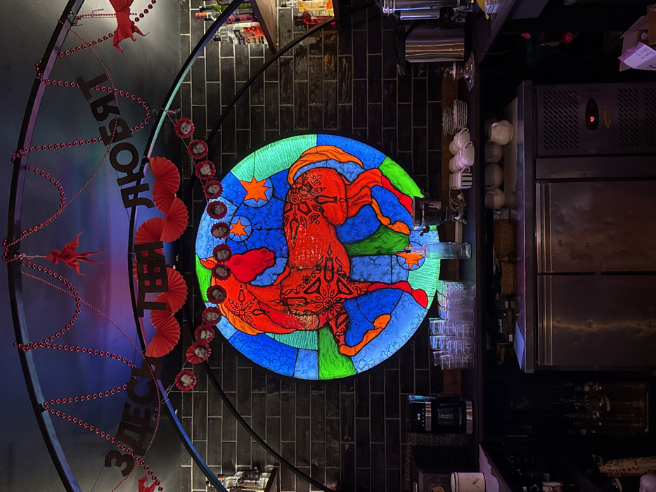 This image depicts an interior view of a bar or cafe in Moscow, Russia. Centered against a dark, segmented masonry wall is a large, circular, backlit artwork styled as stained glass. This artwork features a stylized red horse adorned with intricate patterns, set against a background of blue and green segments with two prominent orange stars.
Above the artwork, a dark curved metal arch serves as a support for various decorations. These include red beaded garlands, red pleated paper fans, and a banner with black Cyrillic letters spelling "ЗДЕСЬ ТЕБЯ ЛЮБЯТ" (Here You Are Loved). Below this banner, a garland composed of multiple circular cutouts, each displaying an individual's face, is strung. Red paper cutouts resembling figures or bows are also interspersed among the upper decorations.
Below the central artwork, a dark-surfaced bar counter holds an assortment of items. From left to right, these include a black drip coffee maker, multiple stacks of clear drinking glasses, a clear glass bottle, a silver traditional samovar, and organized stacks of white ceramic teacups and plates. Beneath the counter are two stainless steel commercial refrigeration units or cabinets. Shelves positioned to the left and right sides of the main artwork display various glass liquor bottles. The scene is dimly lit, with the central artwork serving as a significant light source. No individuals are present within the frame.